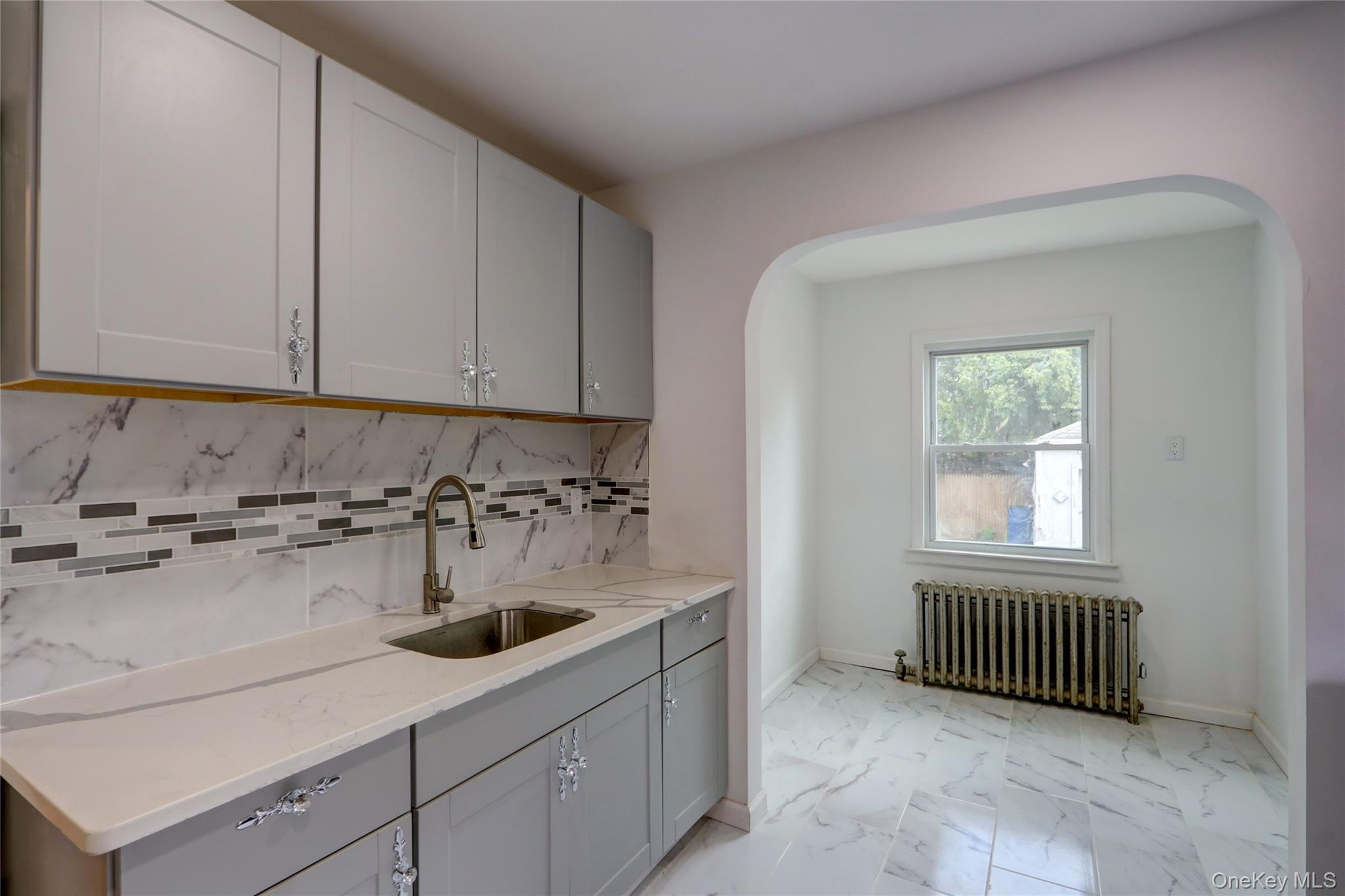 130-55 222nd Street Queens, NY 11413 - Photo 7 of 14 Kitchen featuring backsplash, light marble finish floors, radiator heating unit, and gray cabinetry