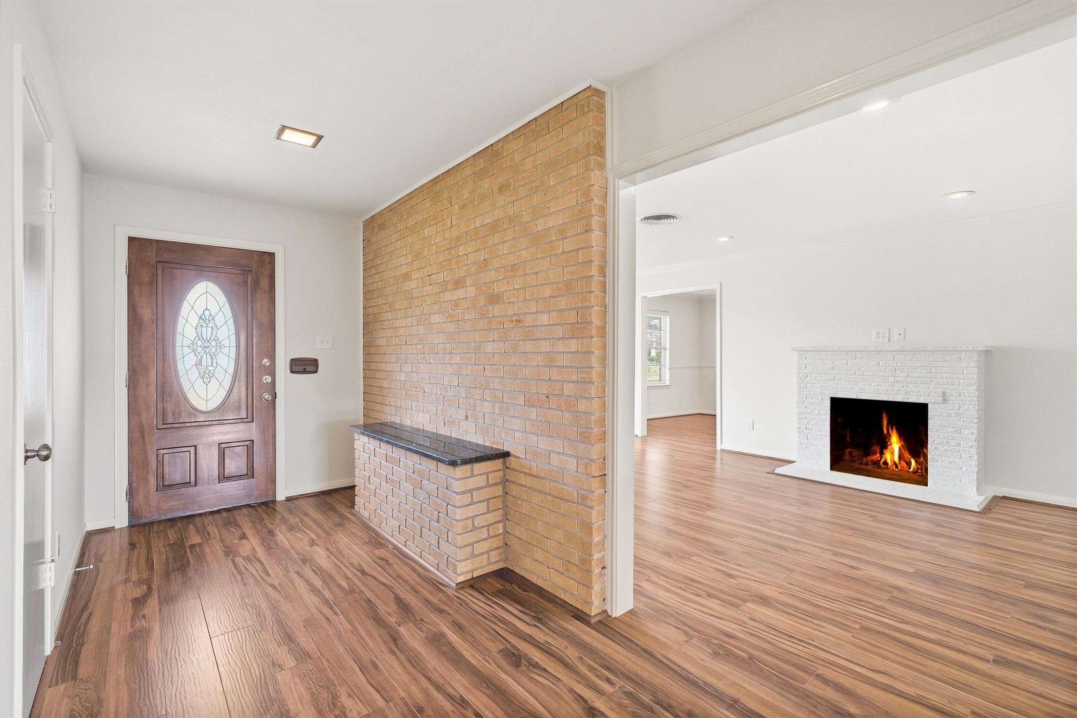 3331 Arbor Street Houston, TX 77004 - Photo 2 of 30 This photo showcases a welcoming entryway with a stylish wood door and brick accent wall. The space flows into a bright living room featuring a cozy, lit fireplace, all set on elegant wood flooring. Ideal for a warm and inviting atmosphere.