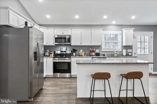 a kitchen with stainless steel appliances granite countertop a stove a sink a refrigerator white cabinets and chairs with wooden floor