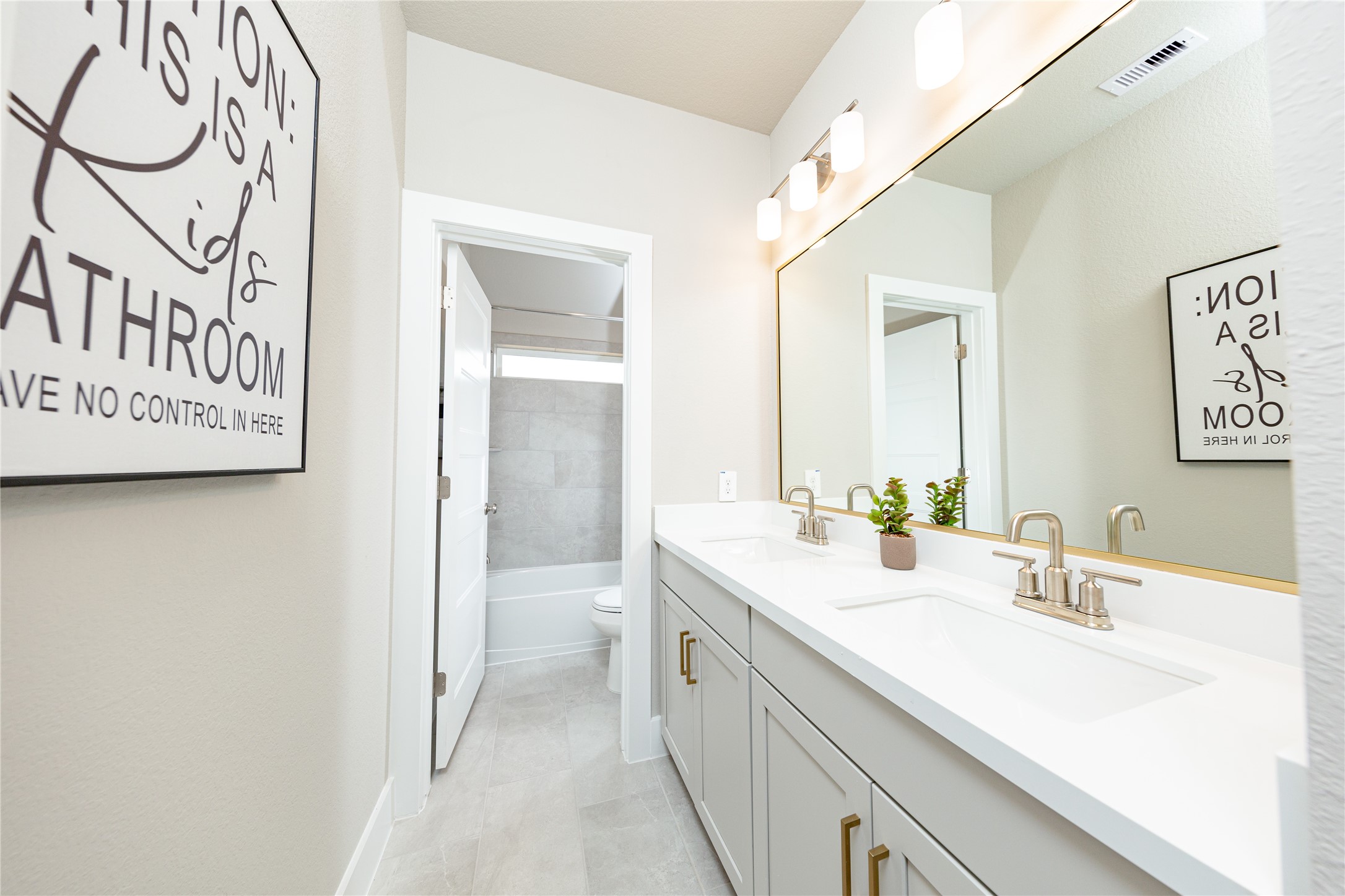 This spacious secondary bathroom features a highly functional layout highlighted by a double-sink vanity with a crisp white countertop and modern cabinetry. An expansive, wall-to-wall mirror reflects the bright overhead lighting, creating an open and airy feel. The thoughtful design includes a separate, semi-private area for the toilet and shower-tub combo, featuring contemporary gray tiling that completes this clean and efficient space.