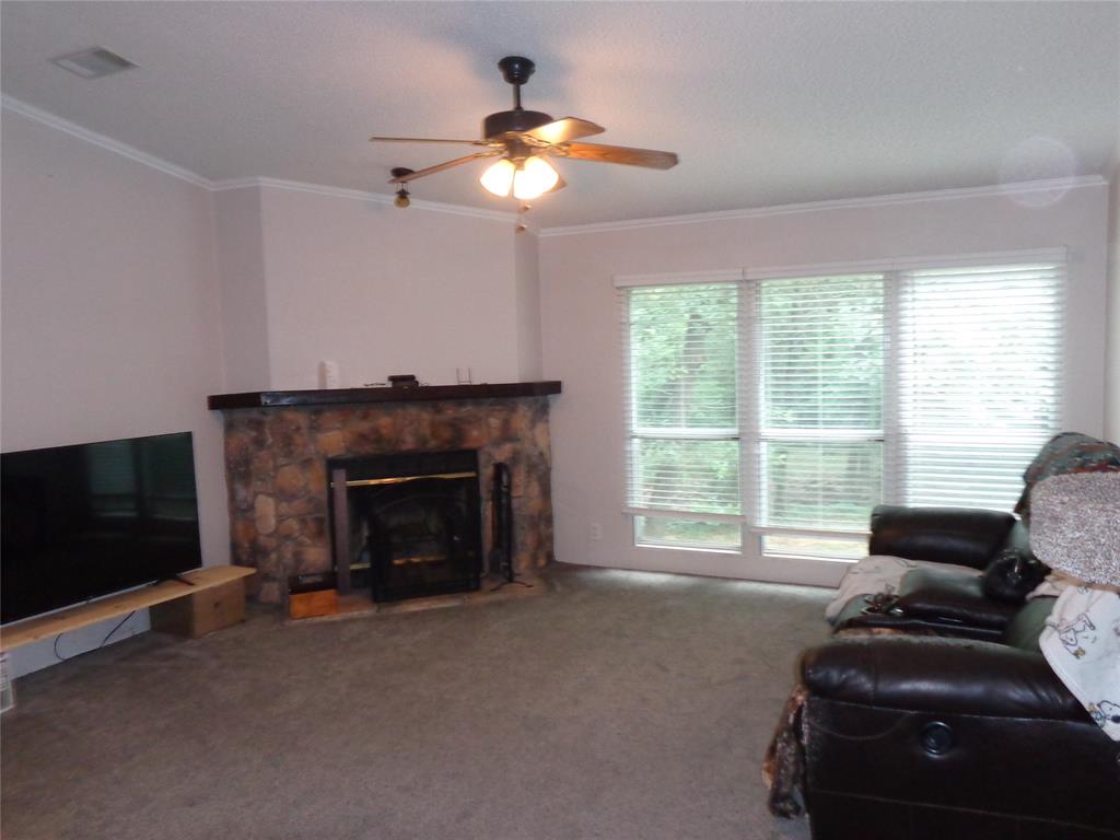 5525 Cedar Oaks Road Grandview, TX 76050 - Photo 13 of 40 Living area featuring a stone-clad fireplace with a dark wood mantel, grey carpeting, a ceiling fan with integrated lighting, and multiple windows with horizontal blinds