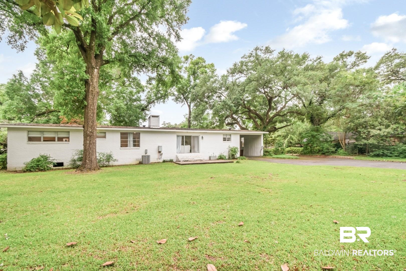 4596 Hawthorne Place Mobile, AL 36608 - Photo 29 of 34 Expansive green lawn with mature trees and a single-story residence featuring white exterior brick, a flat roofline, and a paved driveway leading to a carport