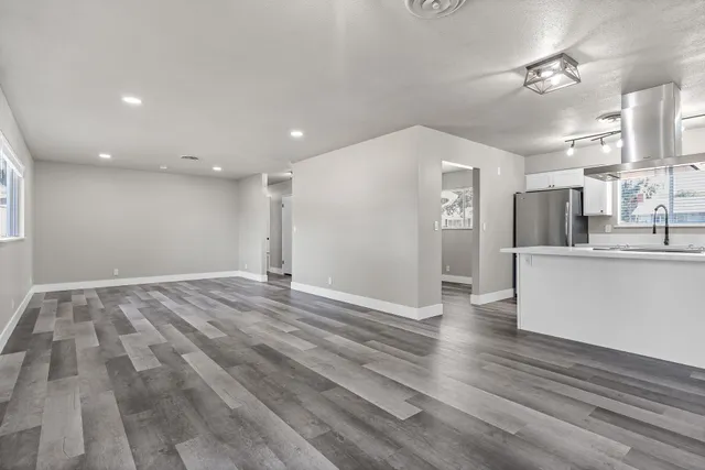 a view of an empty room with kitchen view and wooden floor