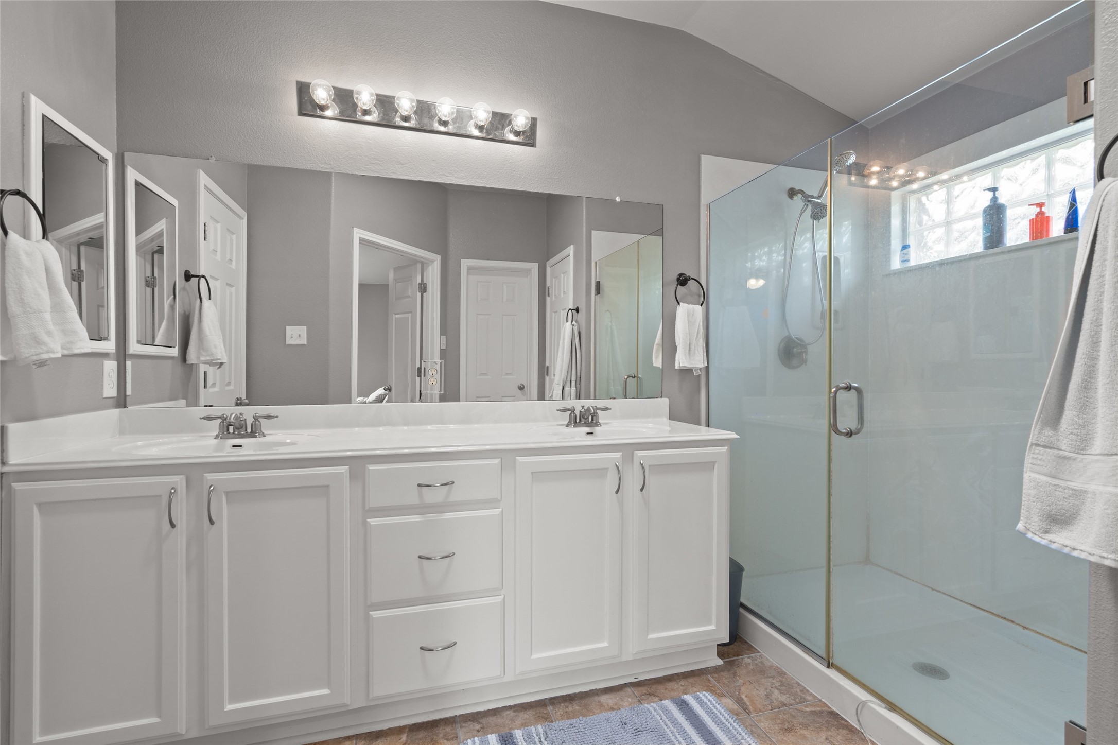 7013 Cromarty Lane Austin, TX 78754 - Photo 22 of 37 The bathroom features a double vanity with white cabinetry, a large mirror, and a glass-enclosed shower with a block glass window