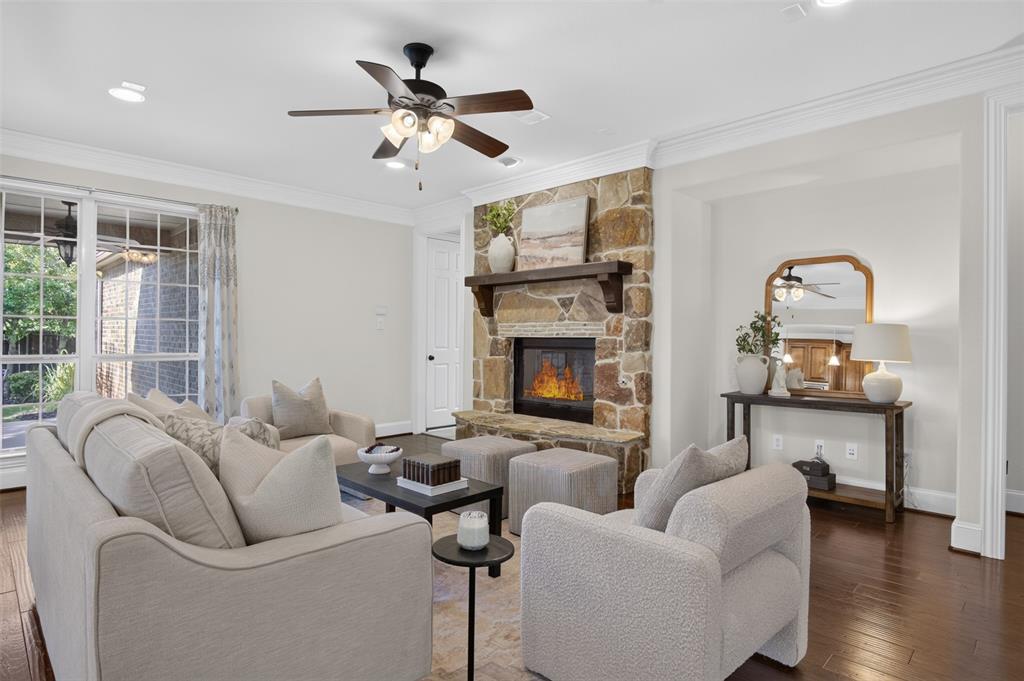 842 Sun Meadow Road Allen, TX 75013 - Photo 9 of 39 Living area featuring a stone-clad fireplace with a wood mantle, rich wood-finish flooring, crown molding, a ceiling fan with integrated lighting, and recessed ceiling lights
