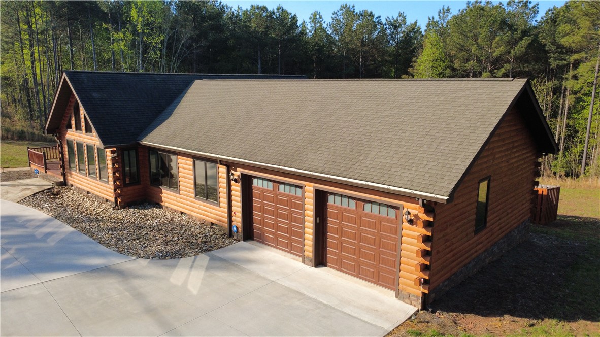 128 McAlister Road West Union, SC 29696 - Photo 36 of 40 A spacious log home with a concrete driveway and an attached two-car garage, set against a backdrop of mature trees.