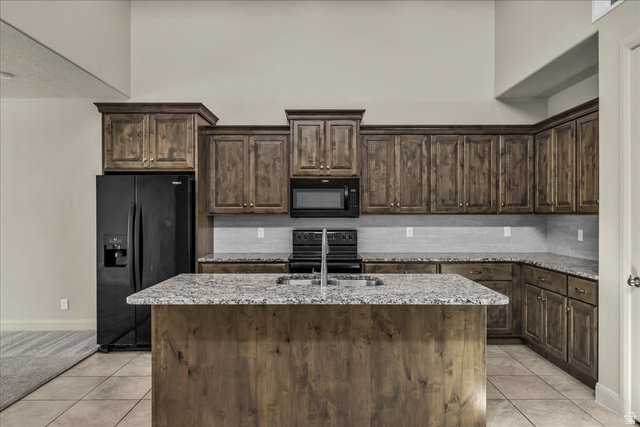 794 North Via Modelo Circle Washington, UT 84780 - Photo 9 of 21 Kitchen featuring dark wood finish cabinets, black appliances, light stone counters, an island with sink, and light tile patterned floors