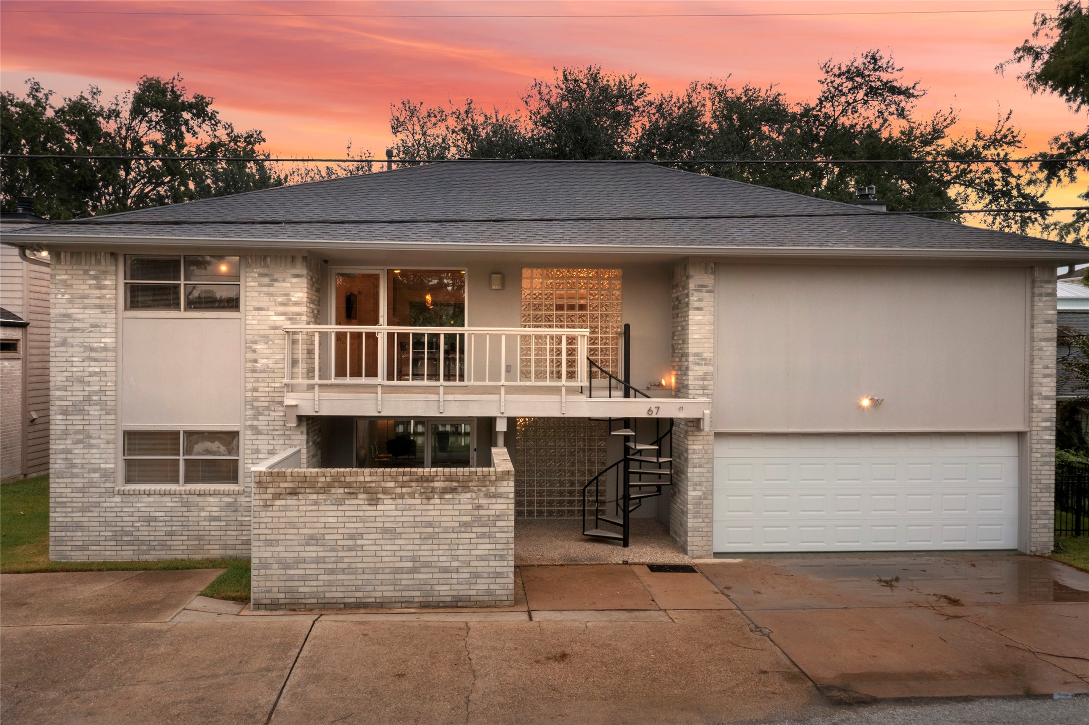 67 Harbor Lane Kemah, TX 77565 - Photo 2 of 47 Discover modern elegance in this two-story home, boasting a sleek design with a spacious balcony, expansive floor to ceiling commercial grade windows, and a striking spiral staircase. The light brick exterior complements a two-car garage, all beautifully illuminated by the warm glow of the setting sun.