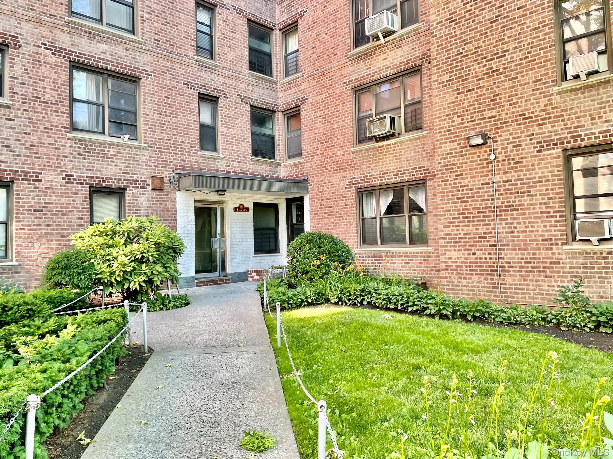 112-25 34th Avenue, Unit 6H Queens, NY 11368 - Photo 46 of 48 Brick facade building featuring a central entry, a concrete walkway, mature shrubbery, and a manicured lawn