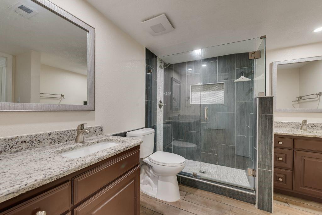 219 West Rochelle Road Irving, TX 75062 - Photo 17 of 29 Bathroom featuring a glass-enclosed shower with dark tile surround, dual vanity areas with granite countertops, and wood-finish flooring