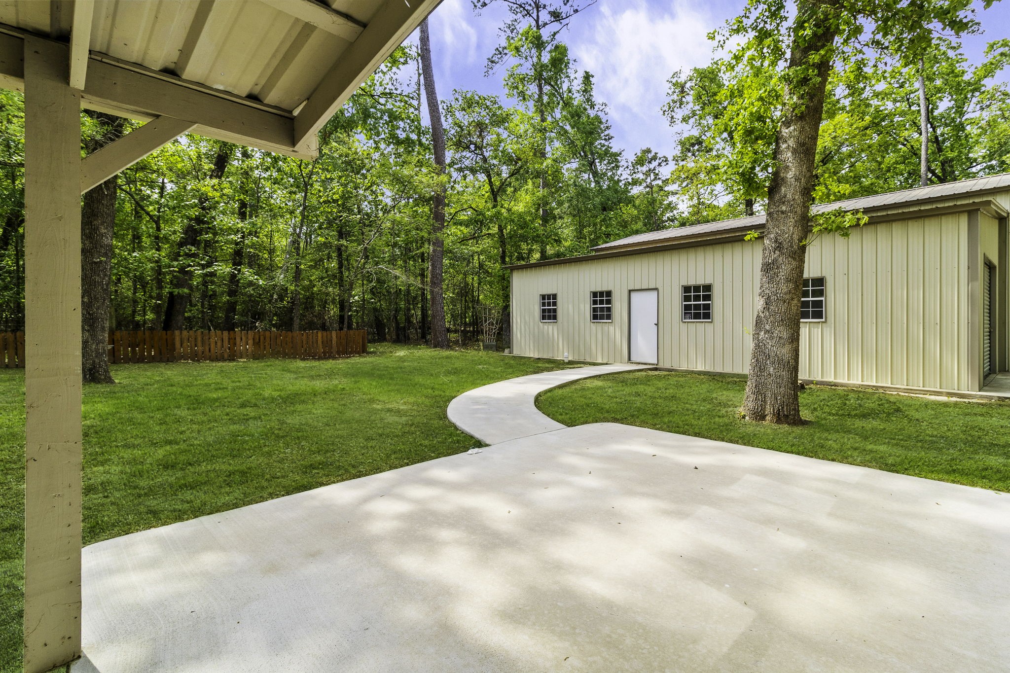 21 Red Oak Circle Point Blank, TX 77364 - Photo 26 of 32 This photo shows a spacious backyard with a paved patio area leading to a large metal outbuilding. The yard features a lush lawn, mature trees, and a wooden fence, offering a serene and private setting.