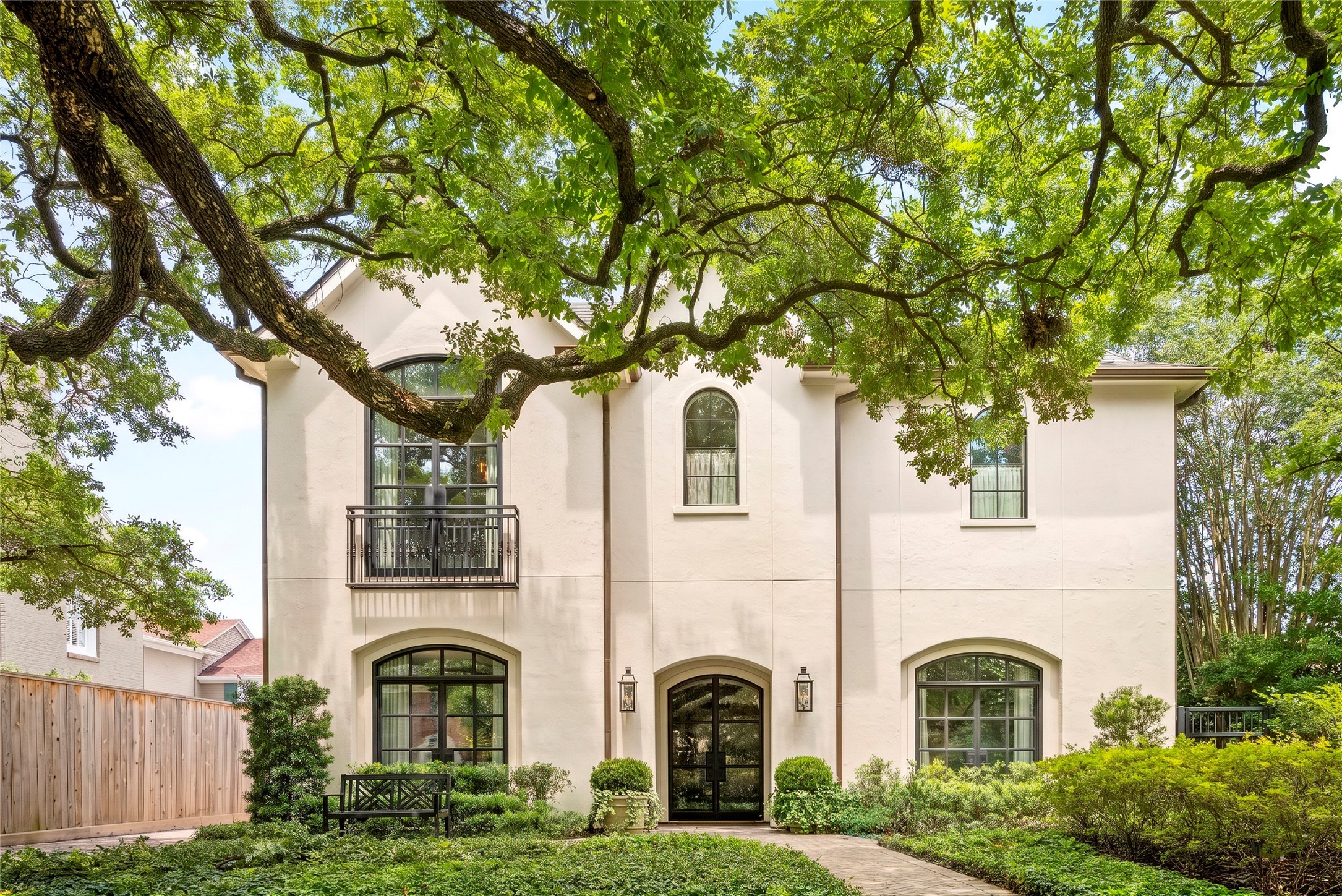 Elegant River Oaks residence framed by mature live oaks, lush landscaping, arched windows, and a gracious stucco façade with a gated entry and balcony detail.