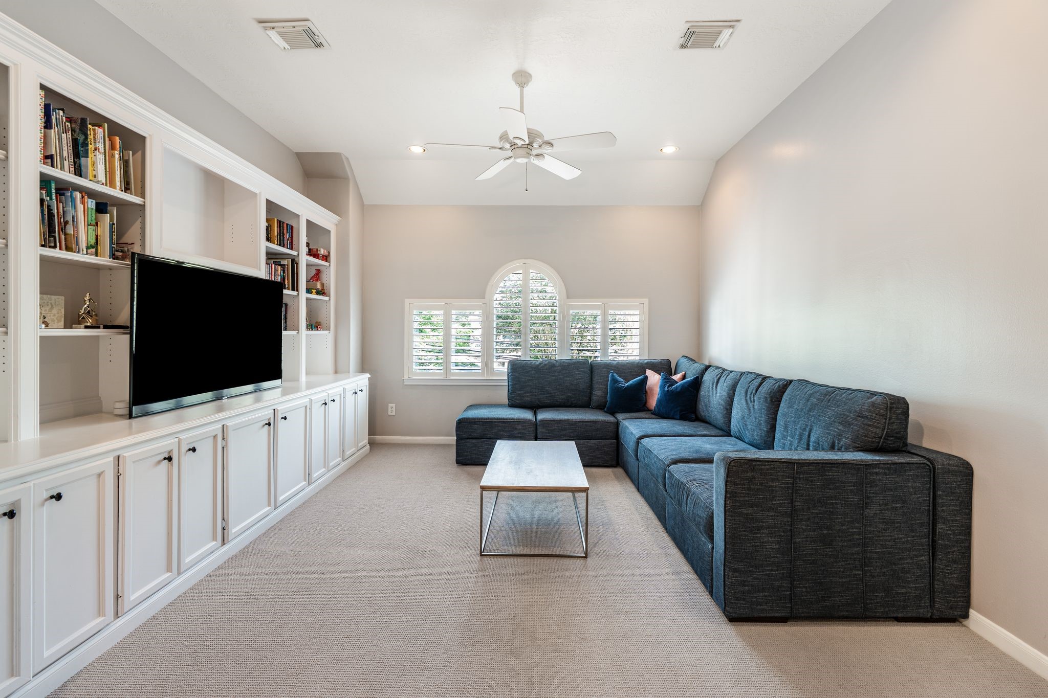 3822 Merrick Street Houston, TX 77025 - Photo 17 of 34 Upstairs, a spacious game room or media room features floor-to-ceiling white built-ins with open shelving, lower cabinet storage, and a dedicated media niche. Carpet flooring, a ceiling fan, and an arched plantation shutter window add comfort and character to this flexible bonus space.