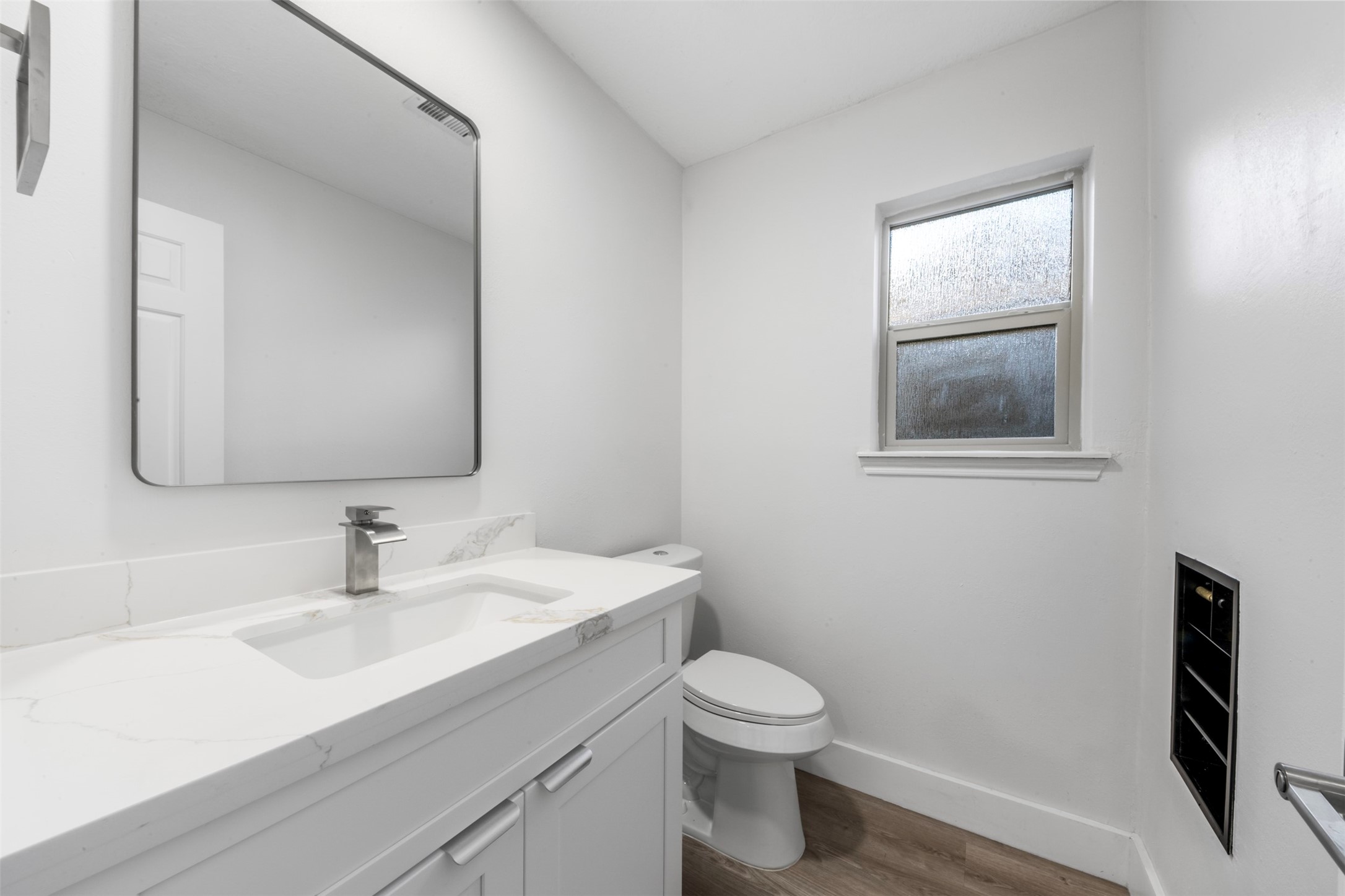 9543 Enstone Circle Spring, TX 77379 - Photo 30 of 50 This is a modern, minimalist bathroom with a sleek white vanity, a large mirror, and a single-handle faucet. It features a toilet, a frosted window for privacy, and wood-style flooring.