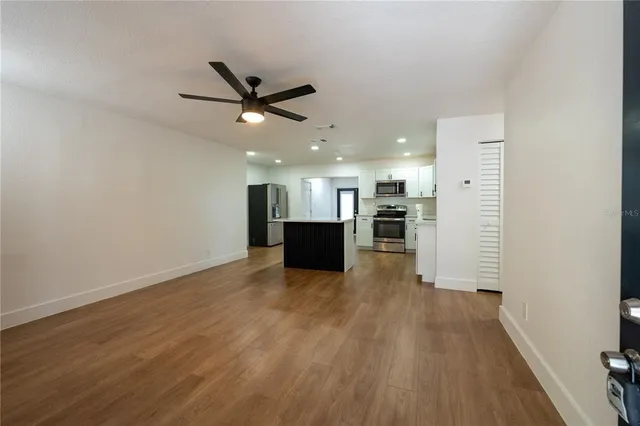 an empty room with wooden floor kitchen view and a ceiling fan
