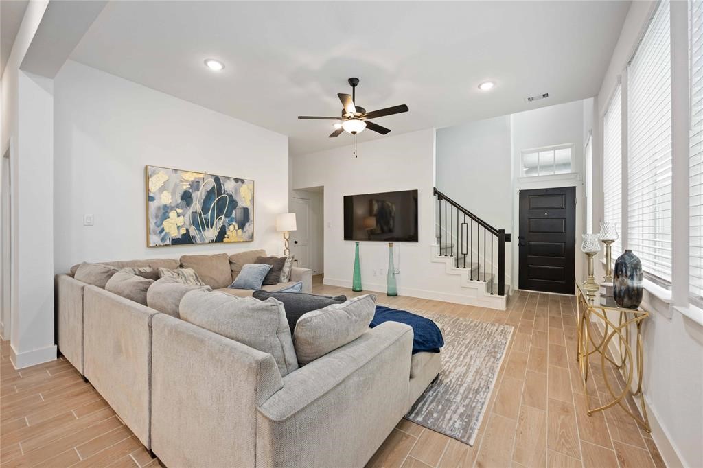 10014 Nectar Path Iowa Colony, TX 77583 - Photo 6 of 26 Spacious living room with modern decor, featuring a large sectional sofa, wall-mounted TV, and stylish ceiling fan. The room has light wood flooring, abundant natural light from large windows, and a staircase leading to the upper level.