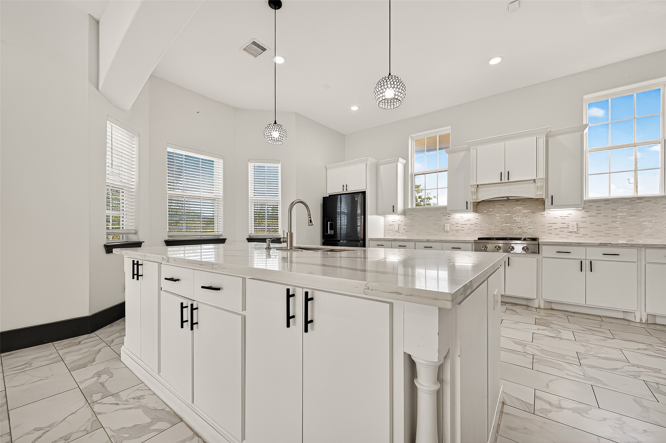 7915 Kirk Road Rosenberg, TX 77471 - Photo 11 of 28 This photo showcases a bright, spacious kitchen with modern white cabinetry and a large marble island. The room features high ceilings, pendant lighting, and ample natural light from multiple windows. The sleek design includes stainless steel appliances and a stylish backsplash, creating a contemporary and inviting space.