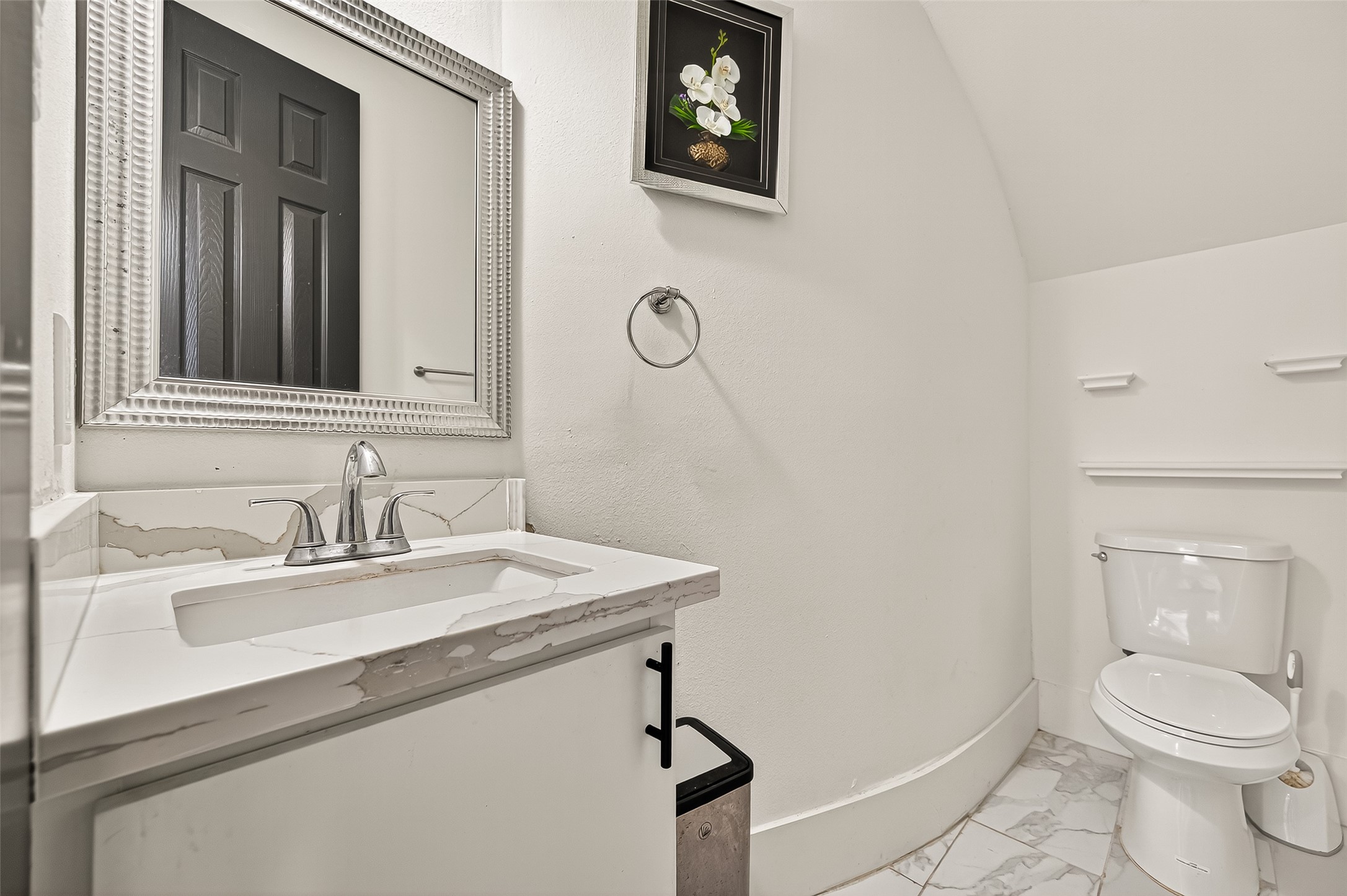 7915 Kirk Road Rosenberg, TX 77471 - Photo 9 of 28 This is a modern bathroom featuring a sleek white vanity with a marble countertop, a large decorative mirror, and a toilet. The room is accented with a floral artwork and has elegant tile flooring.