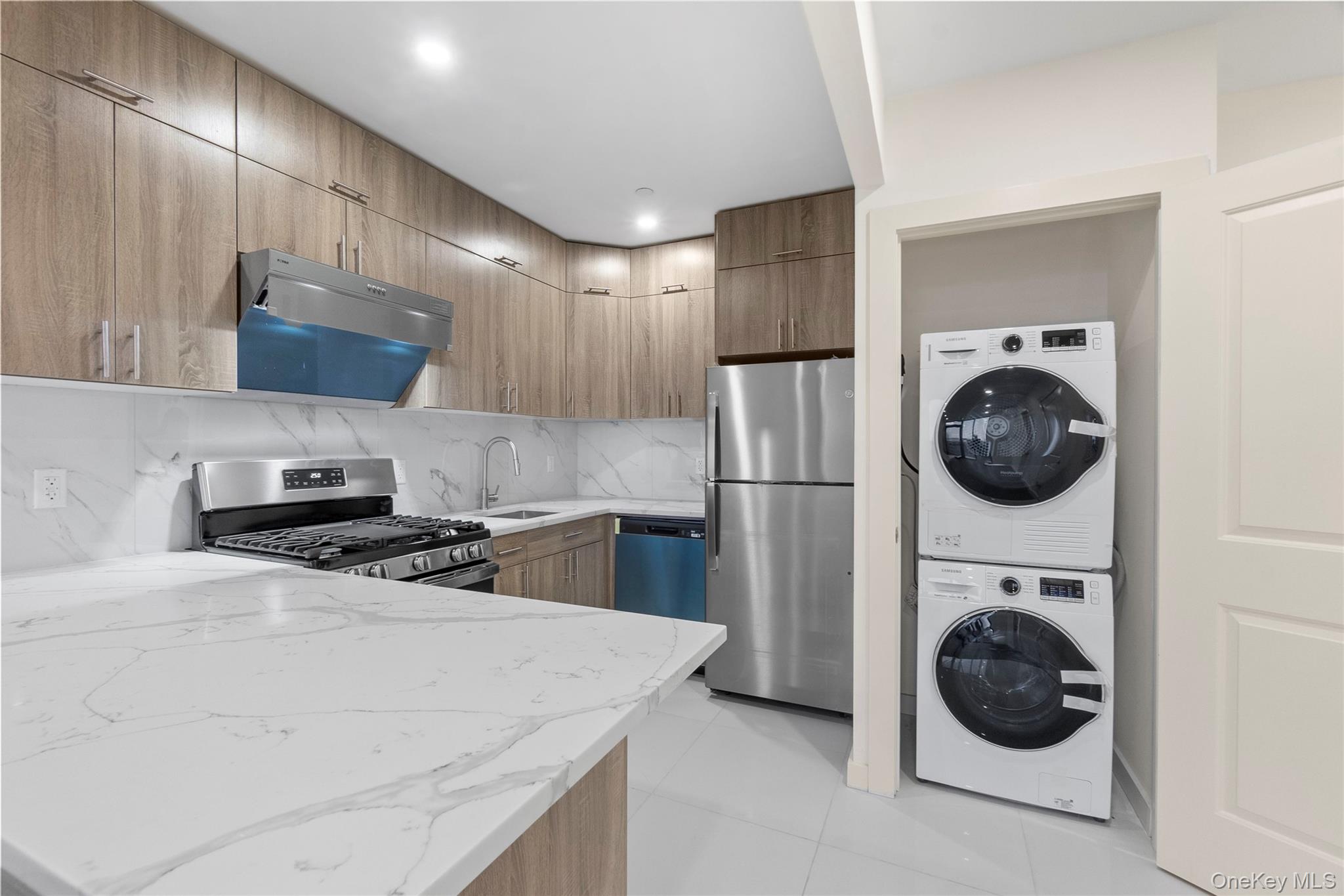 124-28 Queens Boulevard, Unit 12C Queens, NY 11415 - Photo 7 of 14 Kitchen with appliances with stainless steel finishes, light stone countertops, range hood, stacked washer and clothes dryer, and light tile patterned floors