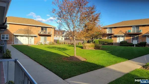 a view of a white house in front of a yard with plants and large tree