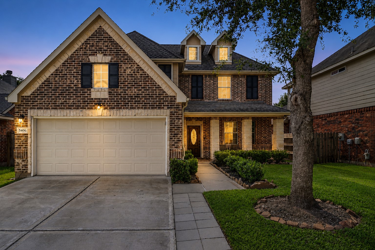 5406 Quarry Ridge Road Richmond, TX 77407 - Photo 2 of 36 Early twilight front exterior highlighting the home's brick elevation, mature shade tree, covered front porch, manicured landscaping, and two-car garage with warm interior lighting creating inviting evening curb appeal.