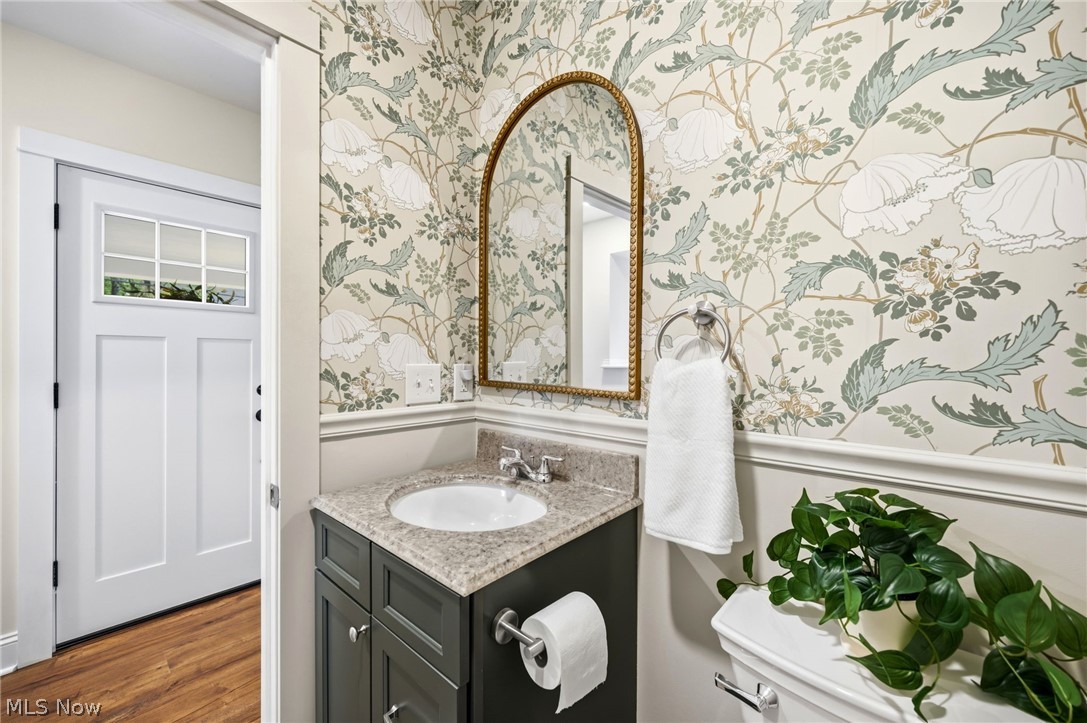 272 Miles Road Chagrin Falls, OH 44022 - Photo 16 of 36 Powder room featuring botanical wallpaper, a dark vanity cabinet with a light-colored countertop, an arched mirror with a gold-tone frame, and wood-finish flooring visible through the doorway