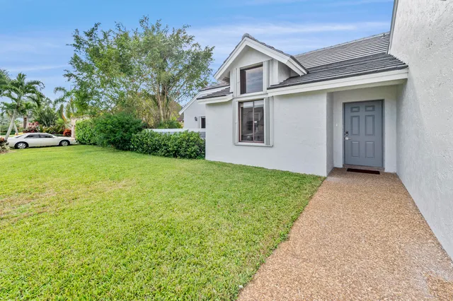 a view of a white house in front of a yard with plants and large tree