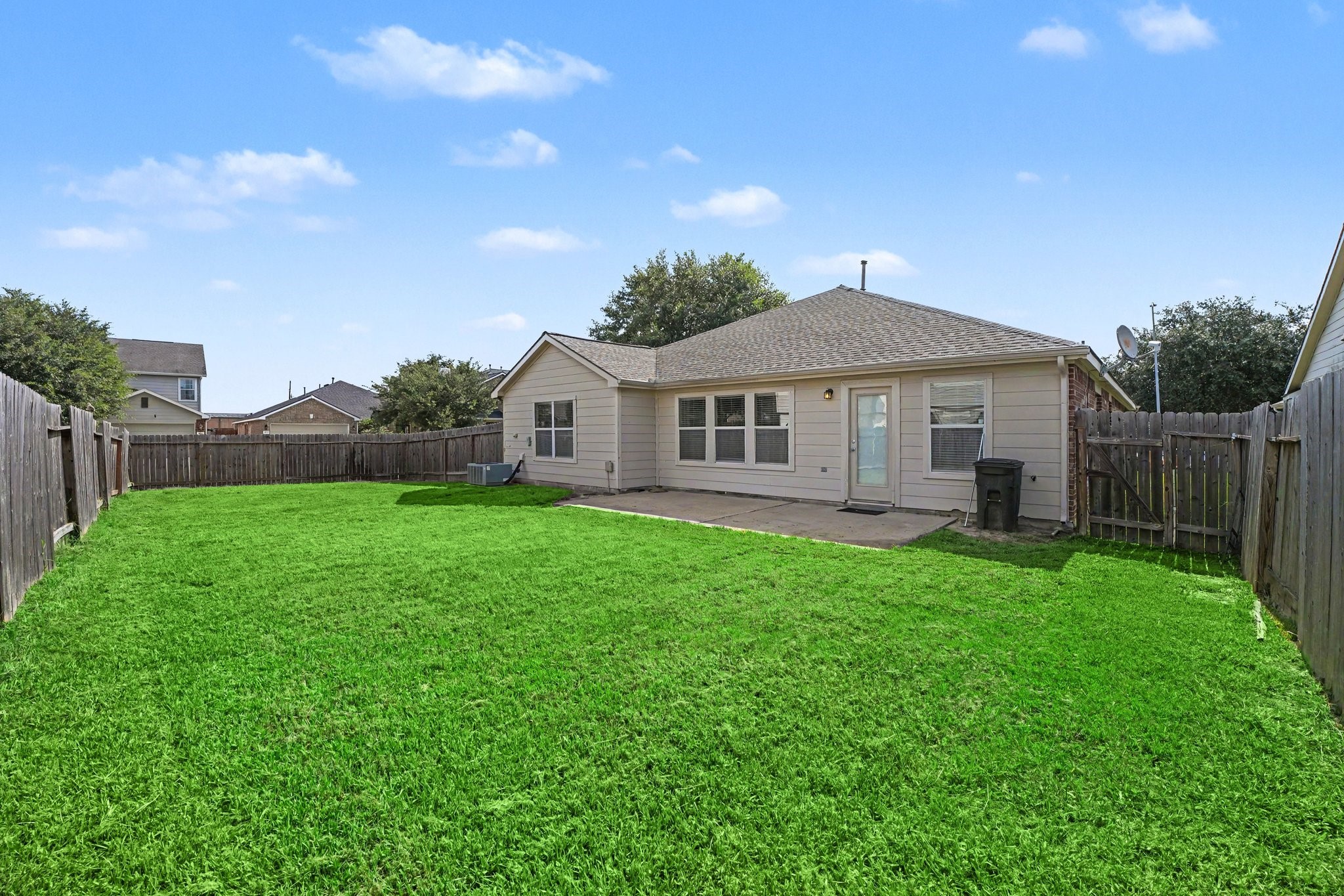 21427 Blooming Rock Lane Spring, TX 77379 - Photo 30 of 32 This photo shows a single-story house with a fenced backyard, featuring a spacious lawn and a concrete patio. The property is surrounded by neighboring homes and has clear skies above.