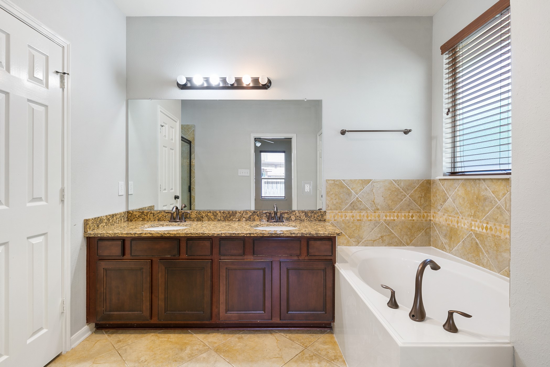 24527 Spring Harbor Drive Spring, TX 77373 - Photo 16 of 22 This bathroom features a double vanity with granite countertops, a large mirror, and stylish lighting. It has a spacious bathtub with elegant bronze fixtures, and the walls are adorned with tiled accents. Natural light enters through a window with wooden blinds, enhancing the inviting atmosphere.