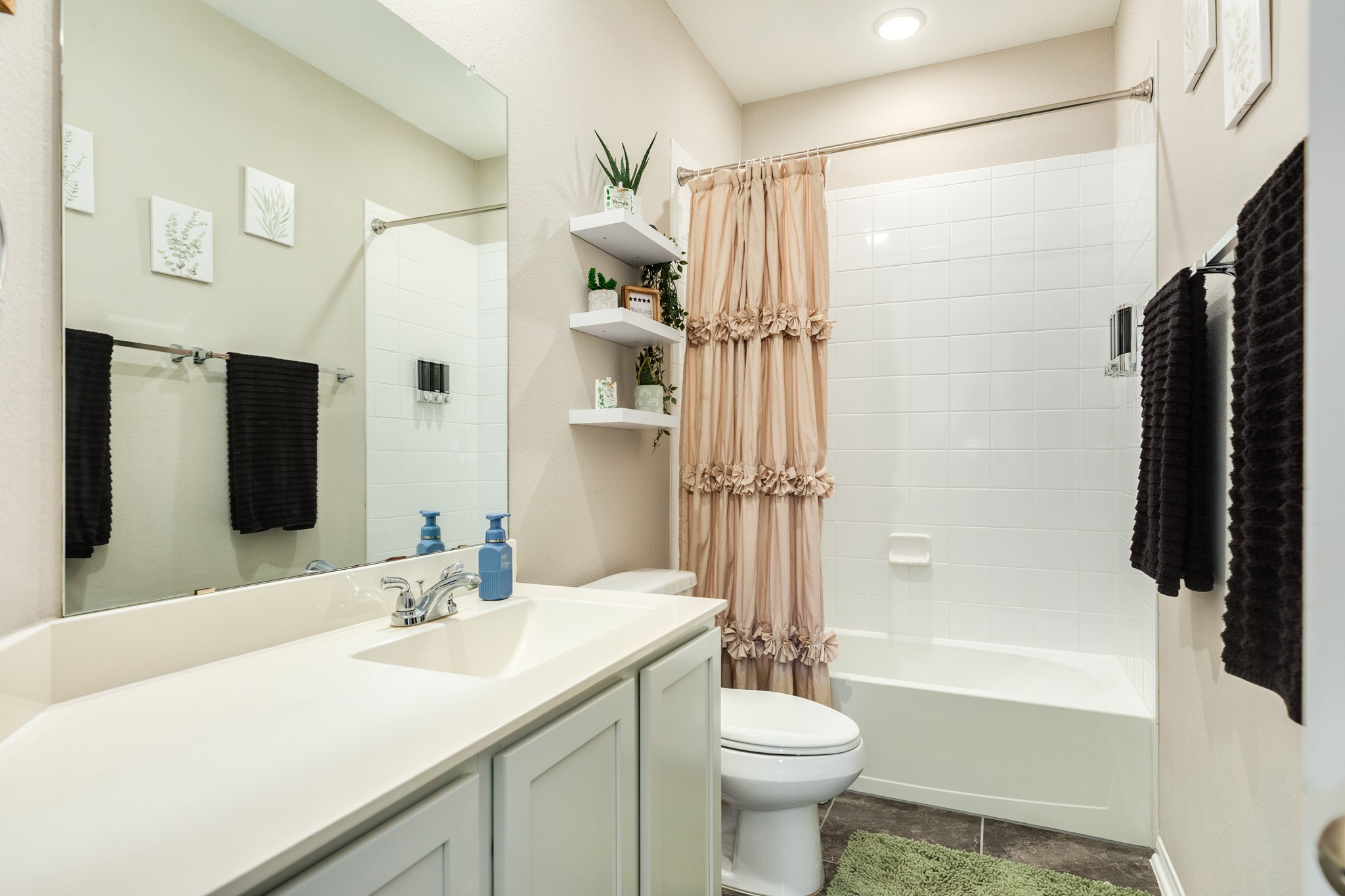 2446 Palisade Crst Drive Rosharon, TX 77583 - Photo 22 of 30 This bathroom features a clean, modern design with a large mirror, white vanity, and built-in shelves. The shower-tub combo has a stylish ruffled curtain, and the space is accented with decorative wall art and plants, offering a fresh and inviting atmosphere.