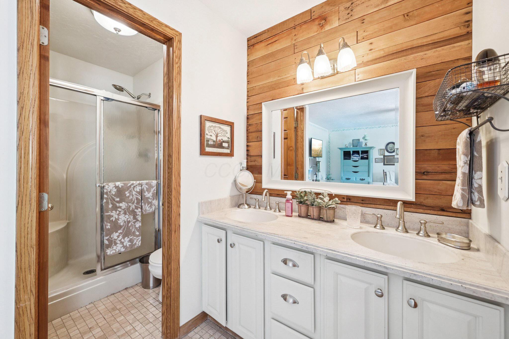 7700 Plumb Road Galena, OH 43021 - Photo 65 of 94 Dual-sink vanity with brushed nickel fixtures, wood accent wall, and spacious mirror adds charm and functionality—plus an adjacent walk-in shower with glass doors and built-in seating.