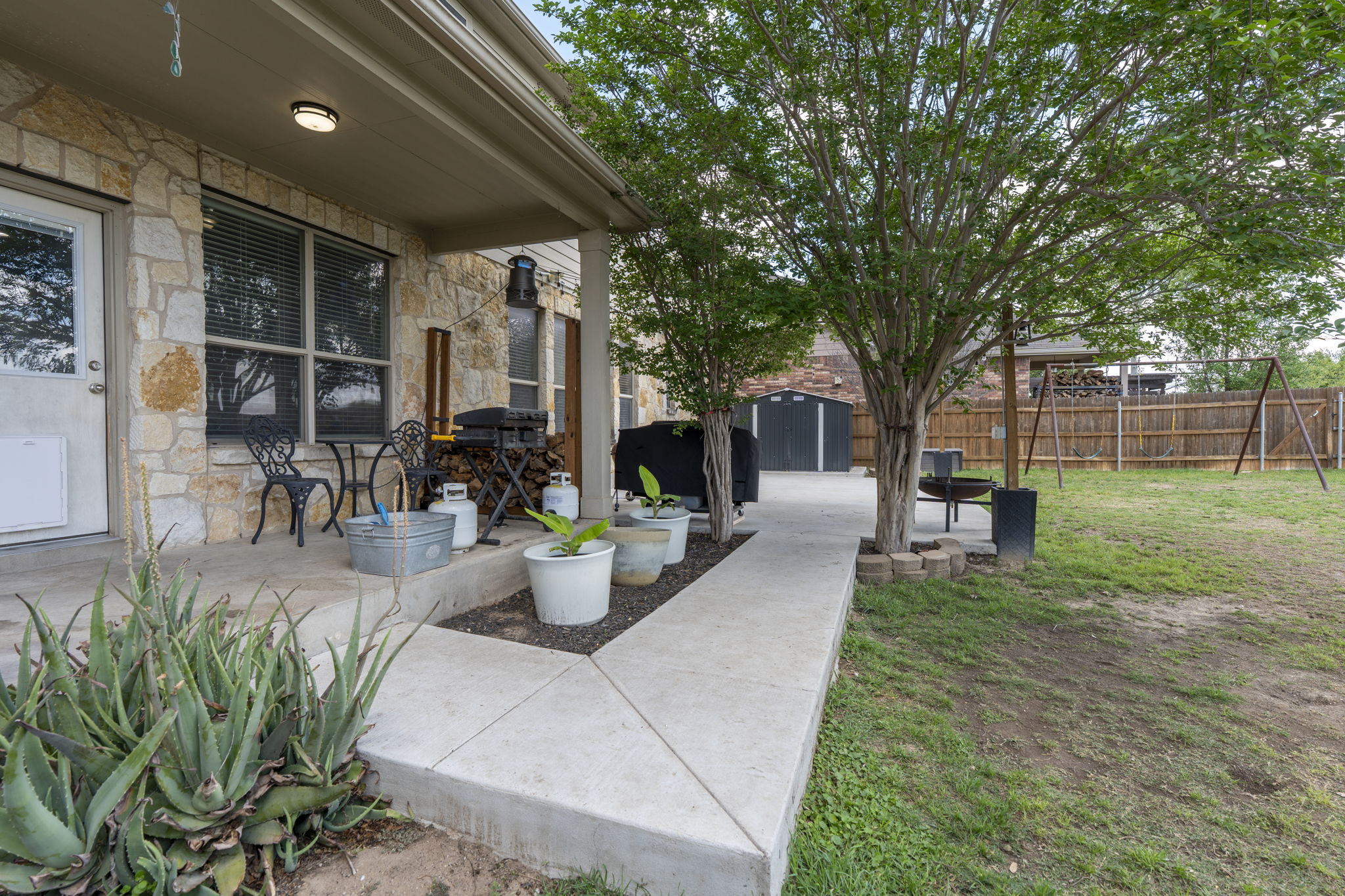227 Sandstone Trail Buda, TX 78610 - Photo 23 of 33 Stone facade exterior with a covered porch, concrete walkway, mature trees, and a wooden fence enclosing the backyard