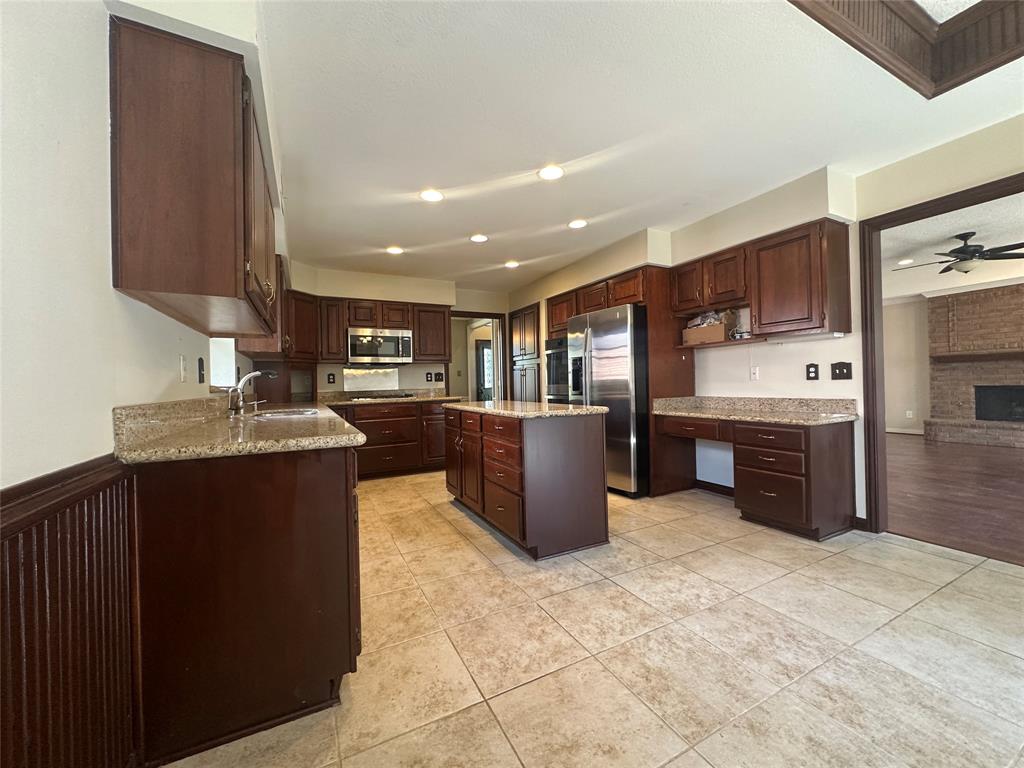 4342 Timberglen Road Dallas, TX 75287 - Photo 13 of 27 Kitchen featuring light stone counters, stainless steel appliances, a kitchen island, light tile patterned floors, and dark wood finish cabinetry