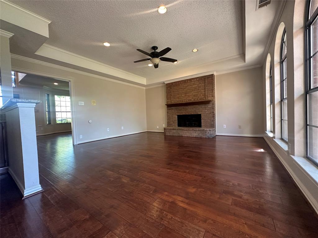 4342 Timberglen Road Dallas, TX 75287 - Photo 6 of 27 Unfurnished living room with a brick fireplace, a tray ceiling, a ceiling fan, ornamental molding, and dark wood-style floors