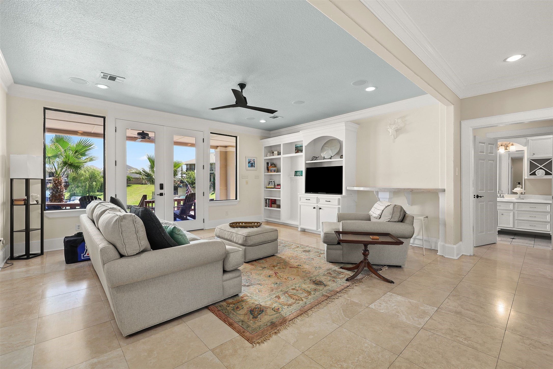 10896 Decatur Street Willis, TX 77318 - Photo 37 of 50 This inviting living room boasts a cozy seating area with a light-colored sofa and armchair, a wall-mounted TV within elegant built-in shelving, large windows flooding the space with natural light and views of the outdoor patio, complemented by tiled flooring, a ceiling fan, and seamless access to a bright bathroom area.