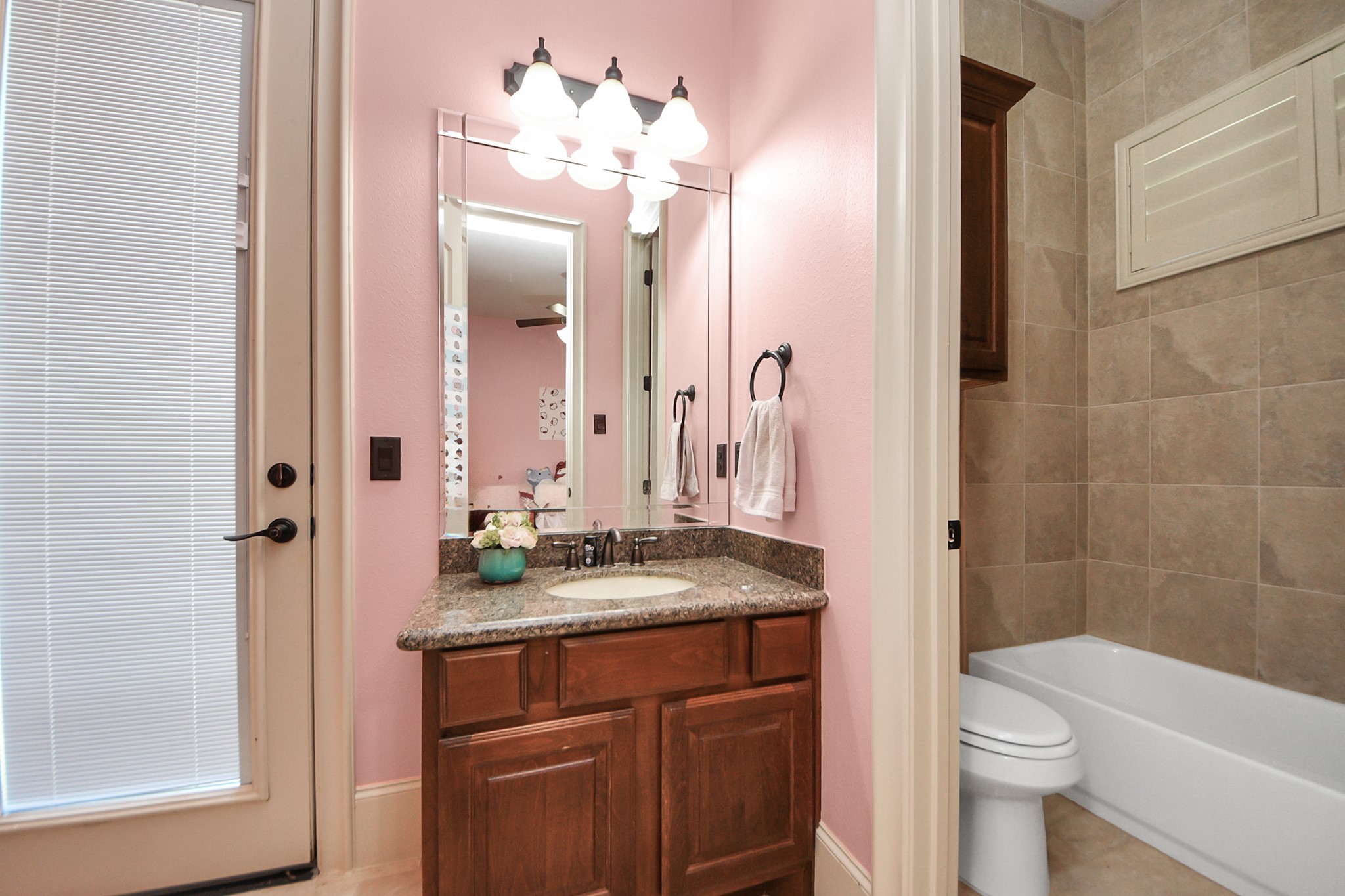 702 Fairway Oaks Conroe, TX 77302 - Photo 30 of 42 This bathroom features a pink
wall with a large mirror and
vanity lighting above a granite
countertop and wooden
cabinetry. It includes a bathtub
with tiled walls and a door with
blinds, offering a charming and
functional space
