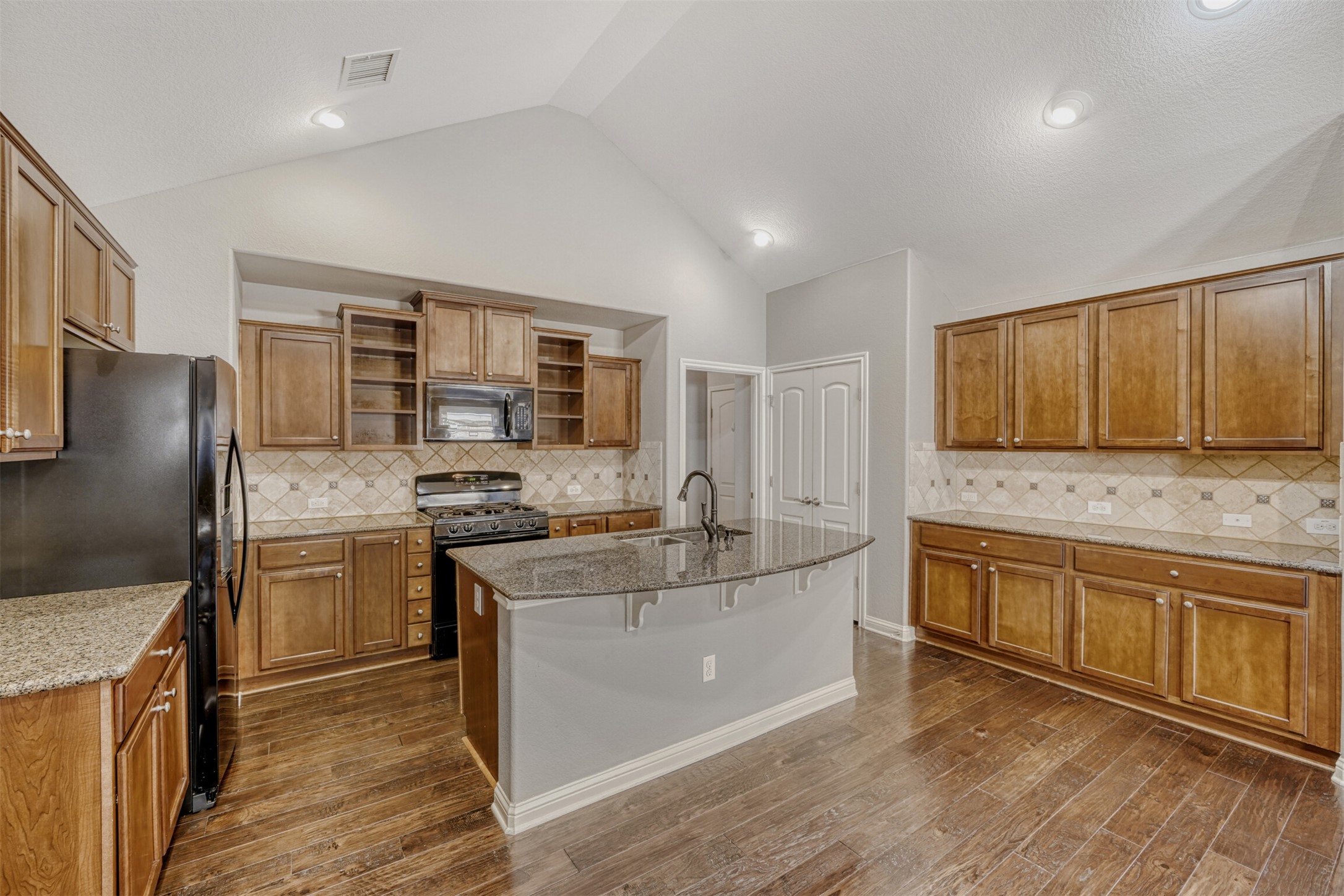 2431 Santa Barbara Loop Round Rock, TX 78665 - Photo 11 of 34 Kitchen with brown cabinets, black appliances, light stone counters, an island with sink, and recessed lighting