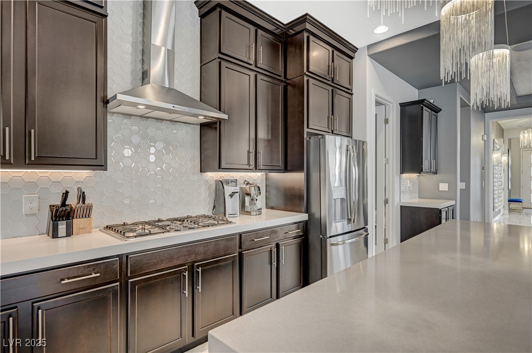 3943 Reyes Avenue Las Vegas, NV 89141 - Photo 17 of 93 Kitchen featuring wall chimney range hood, dark brown cabinetry, decorative backsplash, appliances with stainless steel finishes, and light stone countertops