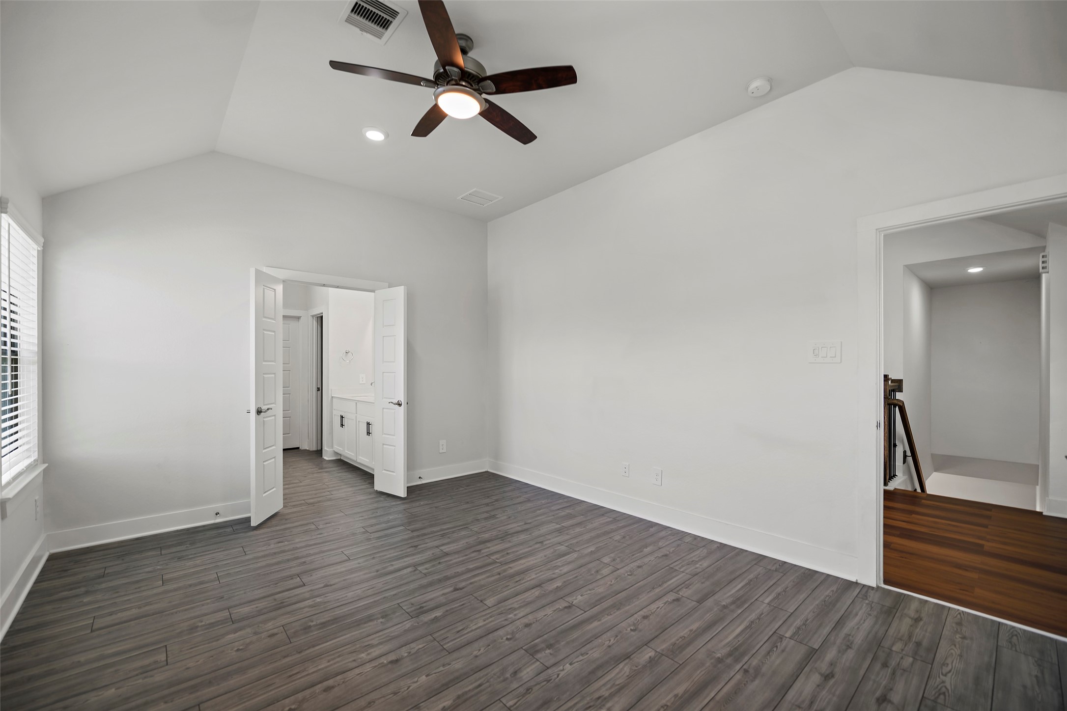 10603 Whitejacket Conroe, TX 77385 - Photo 19 of 42 This spacious Primary Bedroom features modern gray hardwood floors, a vaulted ceiling with a sleek fan, and abundant natural light from a large window. It connects to an en-suite bathroom through double doors, offering convenience and style.