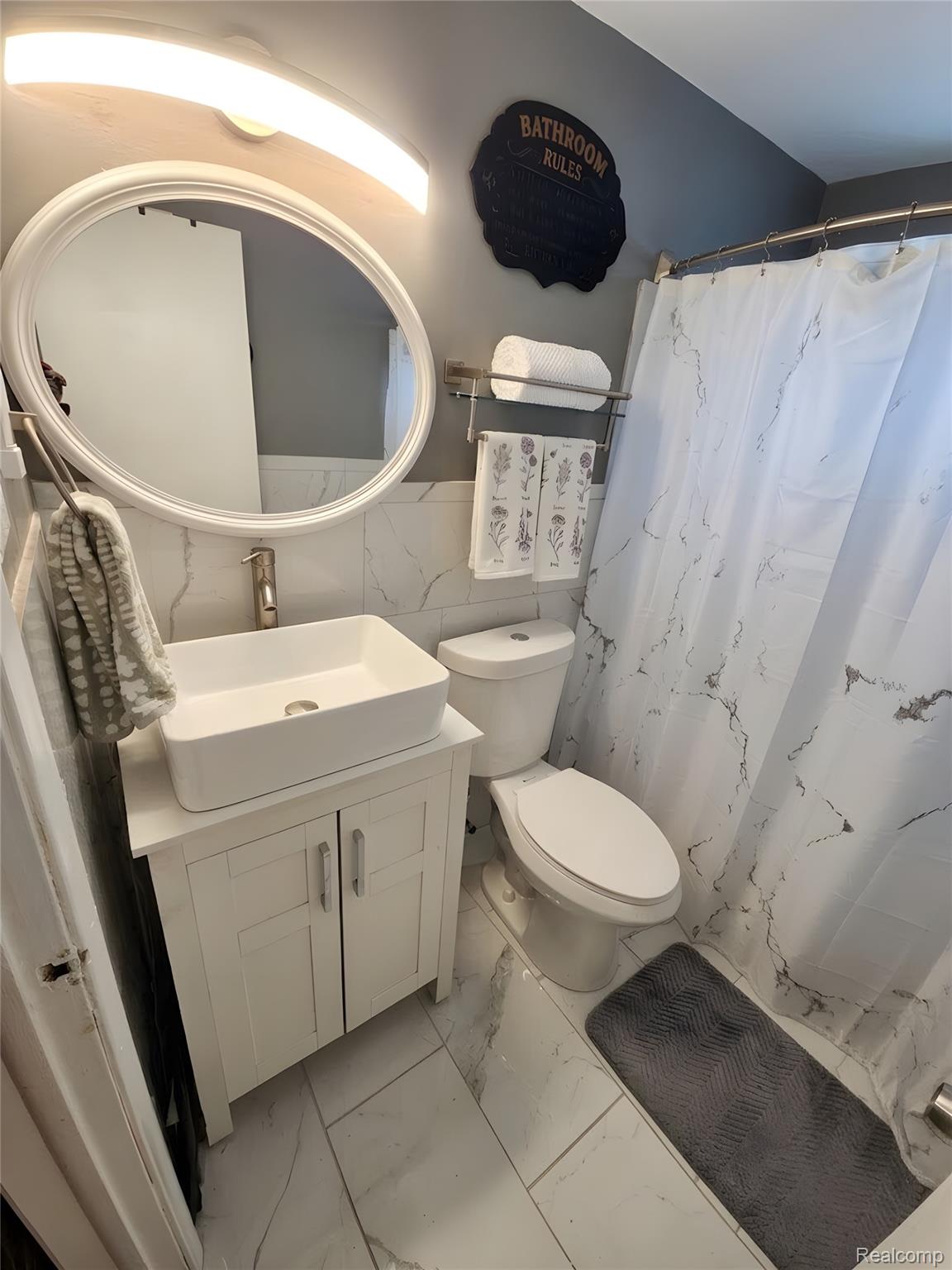 9921 Middlebelt Road, Unit 32 Livonia, MI 48150 - Photo 6 of 11 Bathroom featuring a white vanity with vessel sink, oval mirror, wall-mounted light fixture, and floor-to-ceiling tile in a light tone