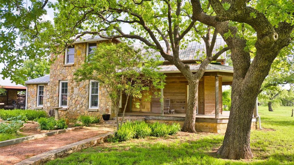 2969 County Lane Talpa, TX 76882 - Photo 11 of 40 Two-story stone and wood-sided residence featuring a wraparound porch, mature trees, a gravel pathway, and lush landscaping