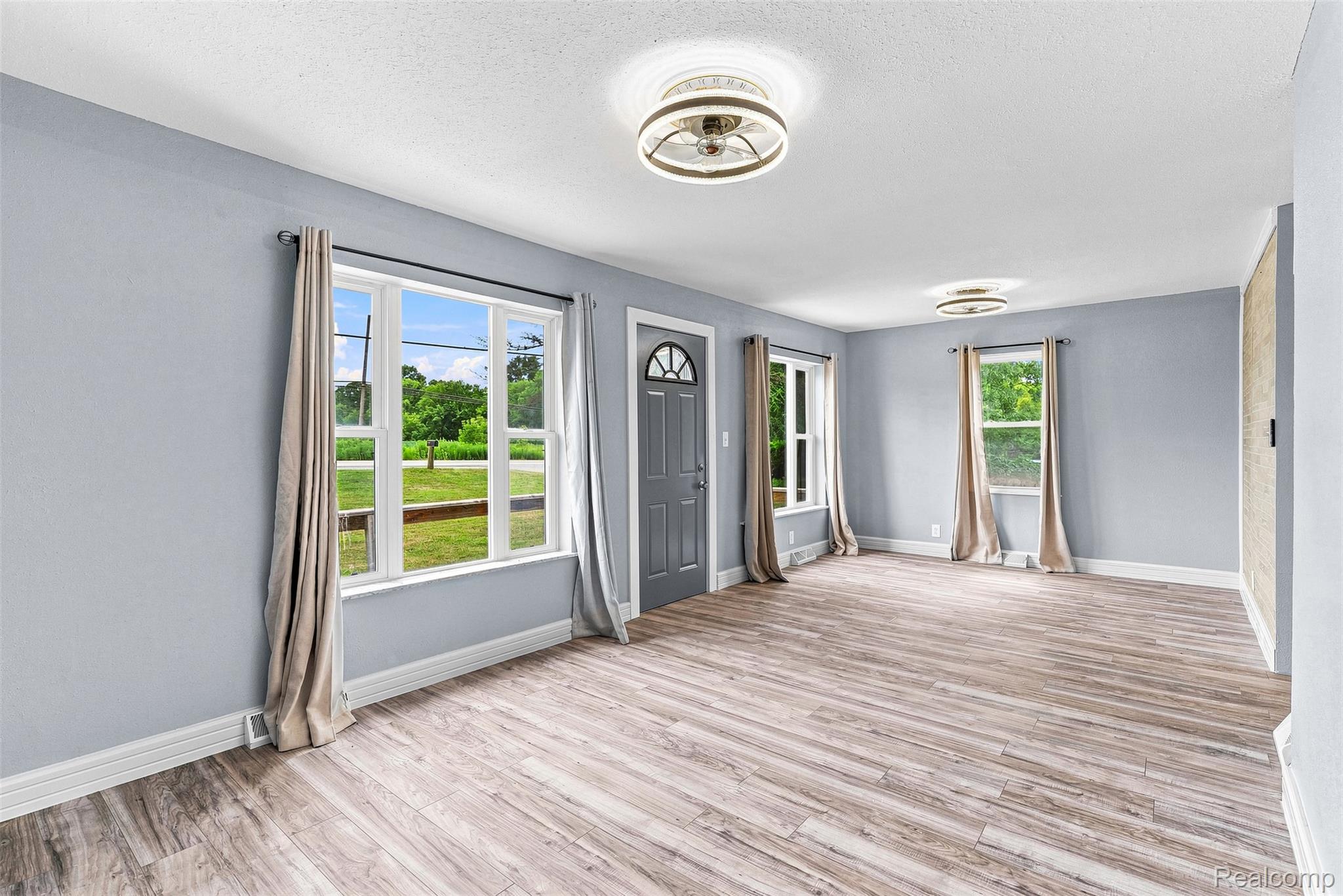 6163 North State Road Davison, MI 48423 - Photo 17 of 45 Spacious room featuring wood-finish flooring, light blue walls, multiple windows, two flush-mount ceiling lights, and a dark-paneled entry door with a decorative glass insert