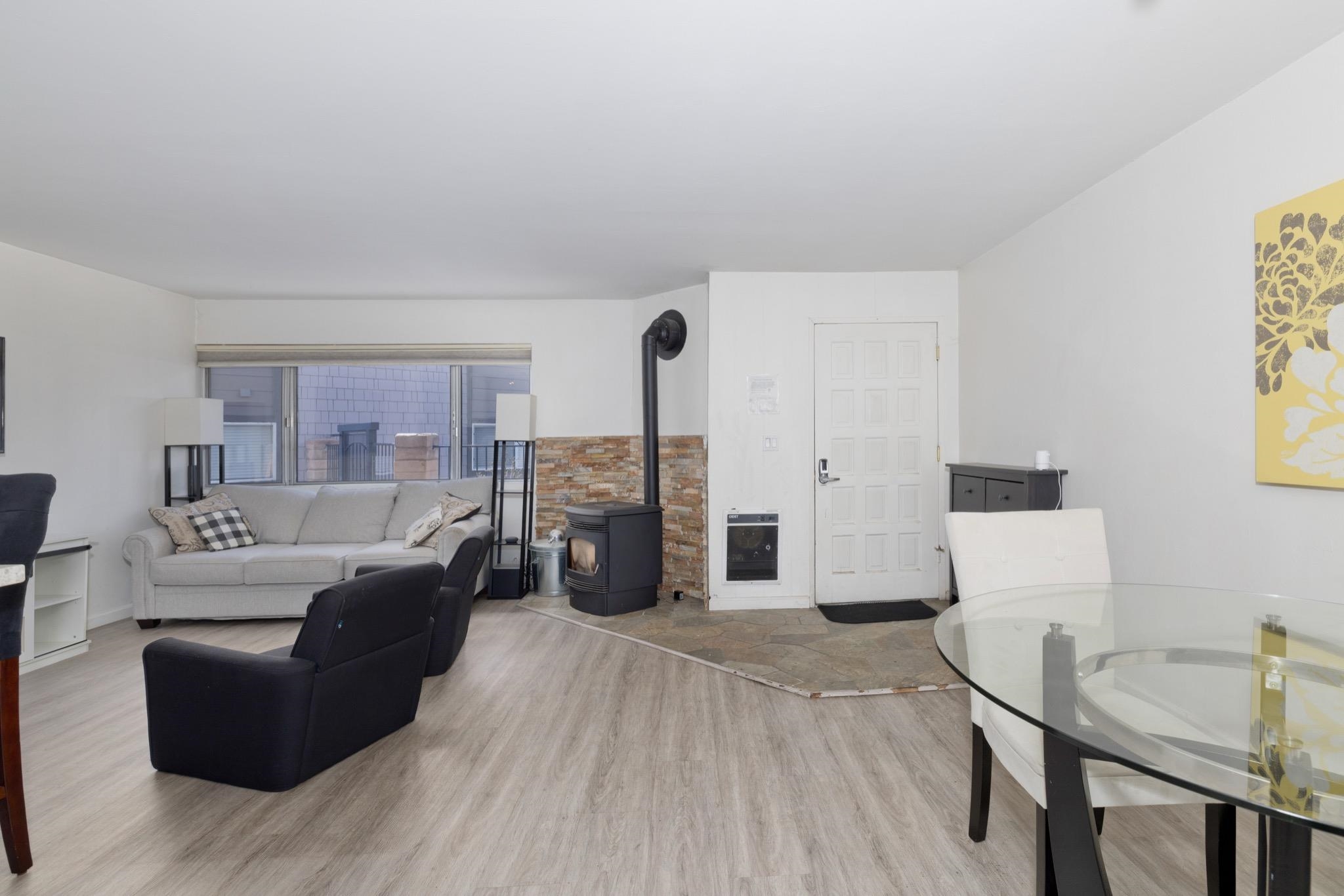 Open-concept living space featuring wood-finish flooring, a contemporary freestanding stove with stone veneer surround, and a large window providing natural light