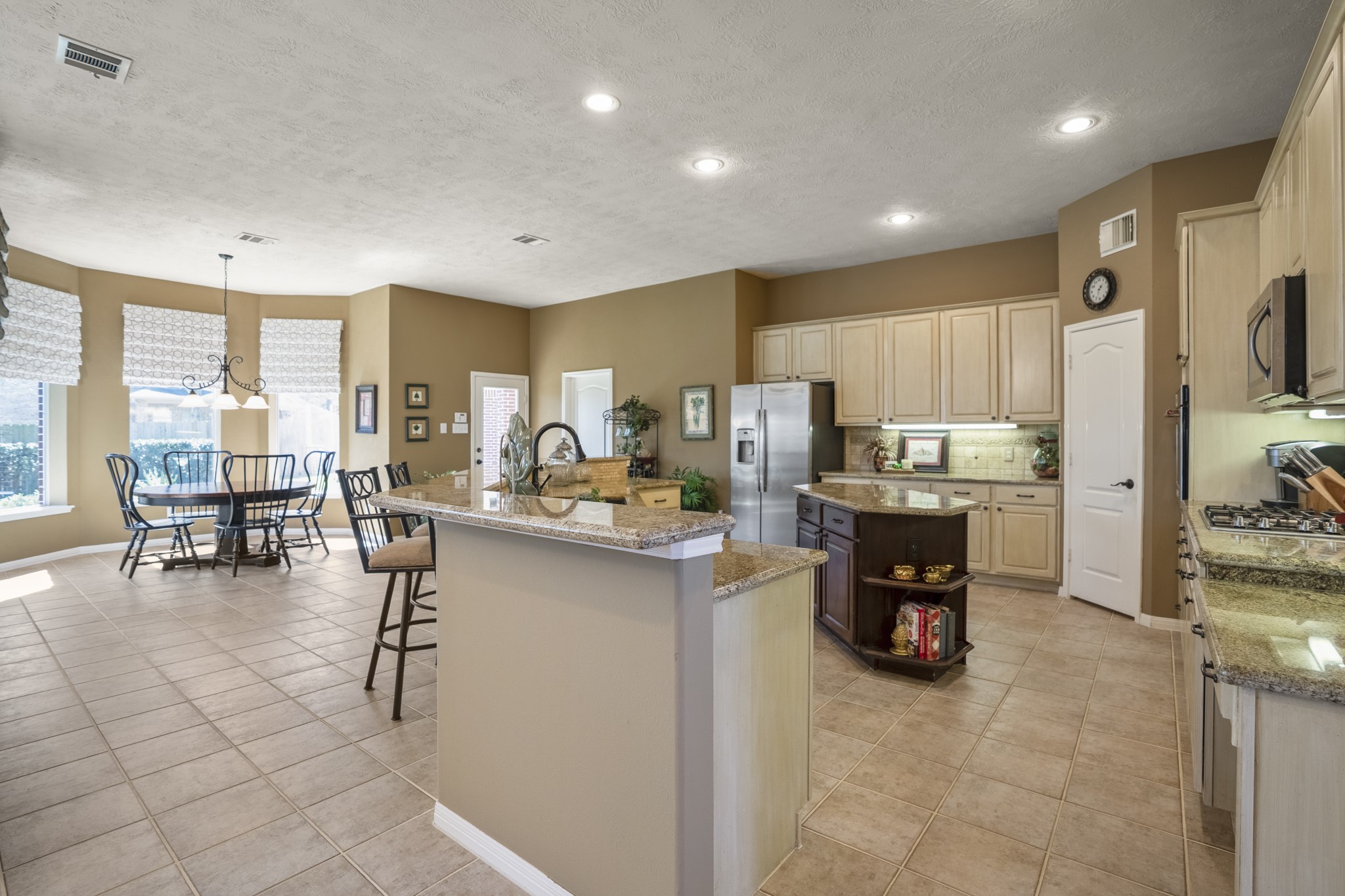 6615 Haughton Drive Spring, TX 77389 - Photo 12 of 50 A closer look at the kitchen reveals a thoughtful two-island layout. The angled island with a raised bar defines the kitchen’s edge, housing the sink while creating an easy spot for casual meals or conversation while dinner comes together.
At the center, a second island floats independently, offering additional storage along with open shelving on either side. Undercabinet lighting softly highlights the countertops, and the gas cooktop with overhead microwave is positioned along the right wall for easy re