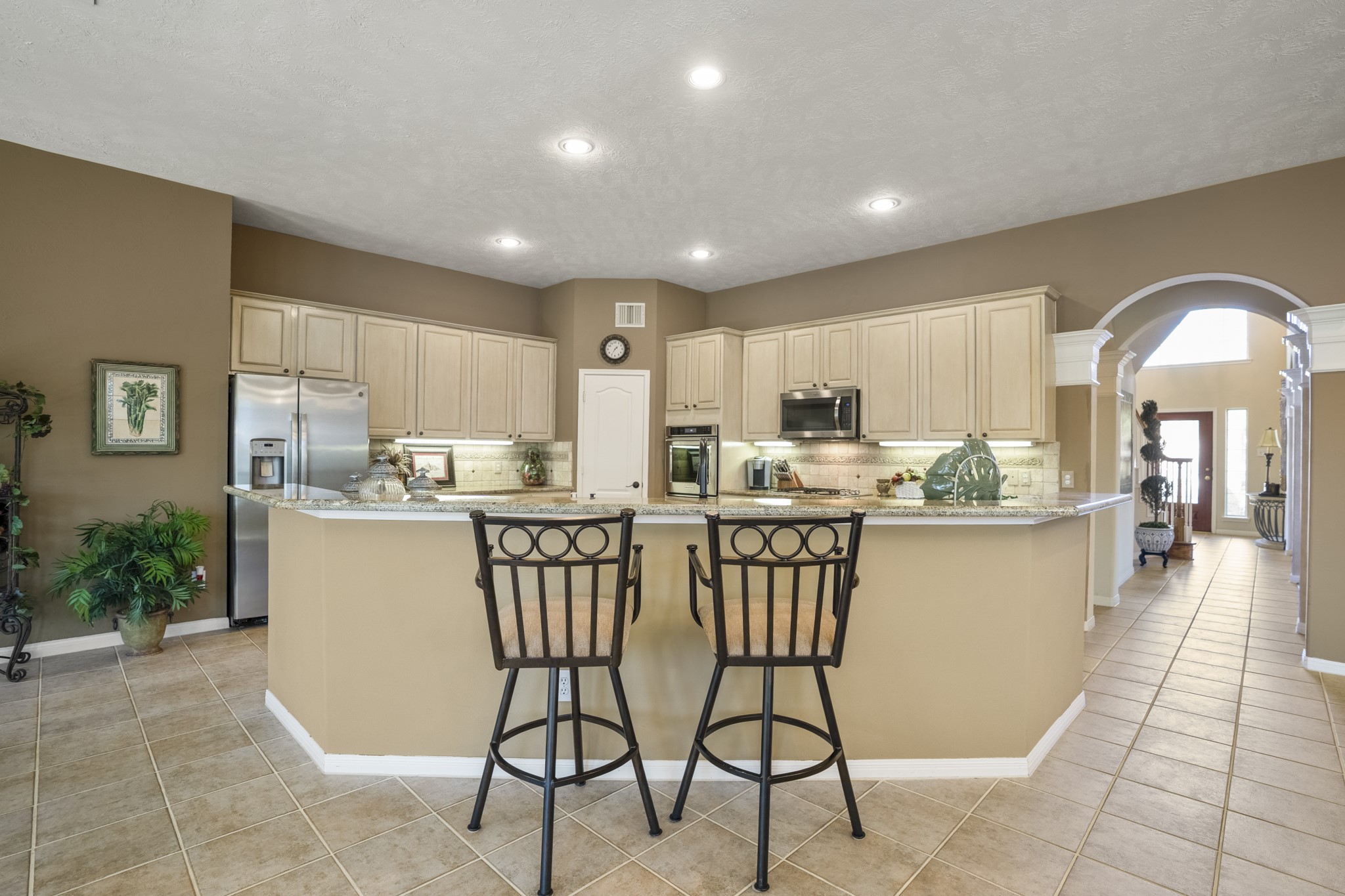 6615 Haughton Drive Spring, TX 77389 - Photo 15 of 50 One last look at the kitchen as a whole. The raised bar island creates a welcoming edge, while the second island adds depth and flexibility at the center. Cabinetry wraps the room with generous storage, stainless appliances shine against the tile backsplash, and sparkling granite counters catch the light from every angle. The built-in desk area near the arched doorway adds a thoughtful touch, keeping daily details within reach without interrupting the design.