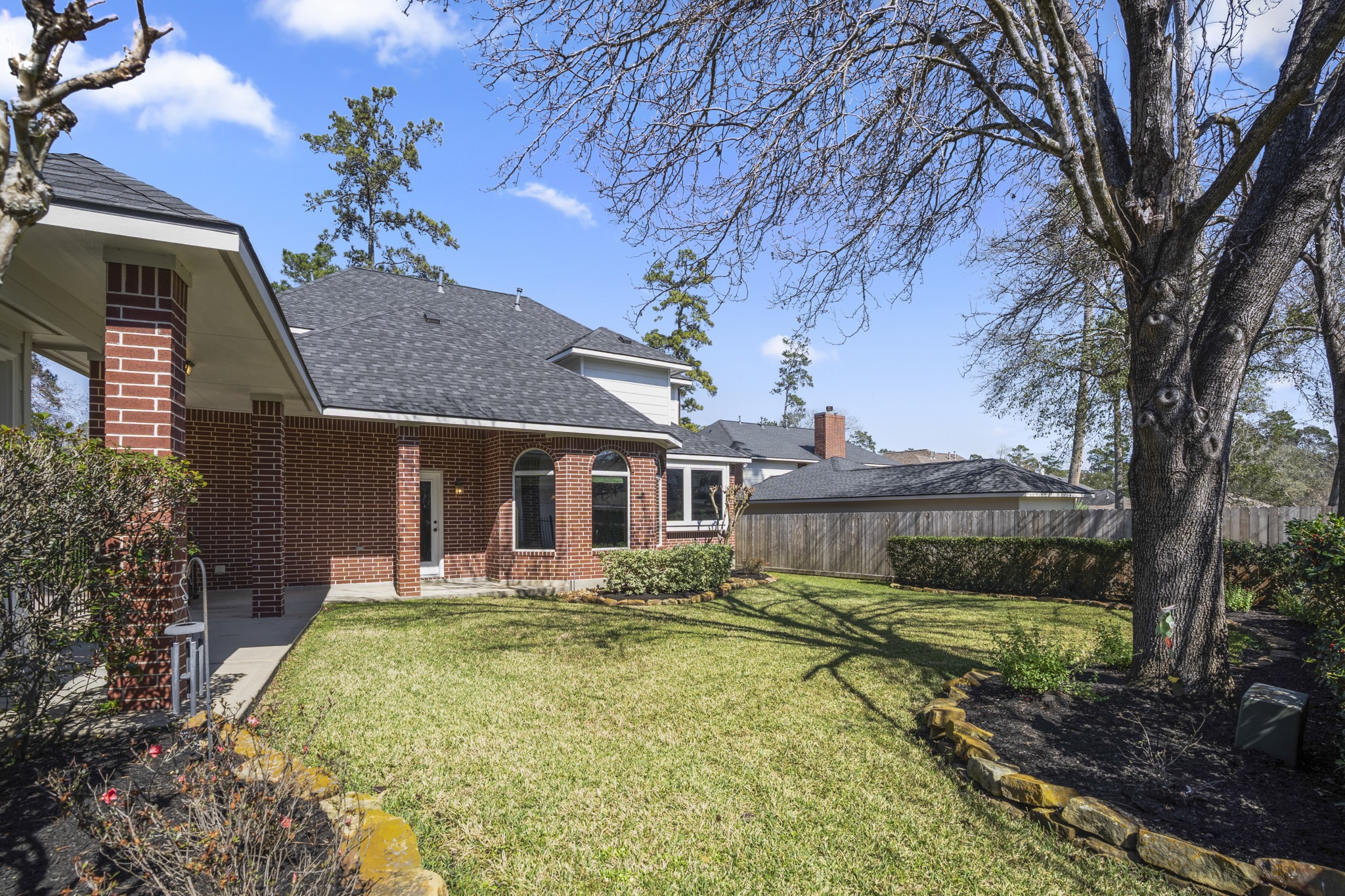 6615 Haughton Drive Spring, TX 77389 - Photo 39 of 50 From this vantage point near the breezeway, the connection between the home and detached garage becomes even more clear, with the covered walkway bridging the two. Just beyond, you catch your first glimpse past the bay windows of the breakfast area, seeing how the yard wraps around the rear of the home. The lawn opens up wider from here, framed by established hedges and mature trees, giving the backyard a sense of space that extends beyond the patio itself.