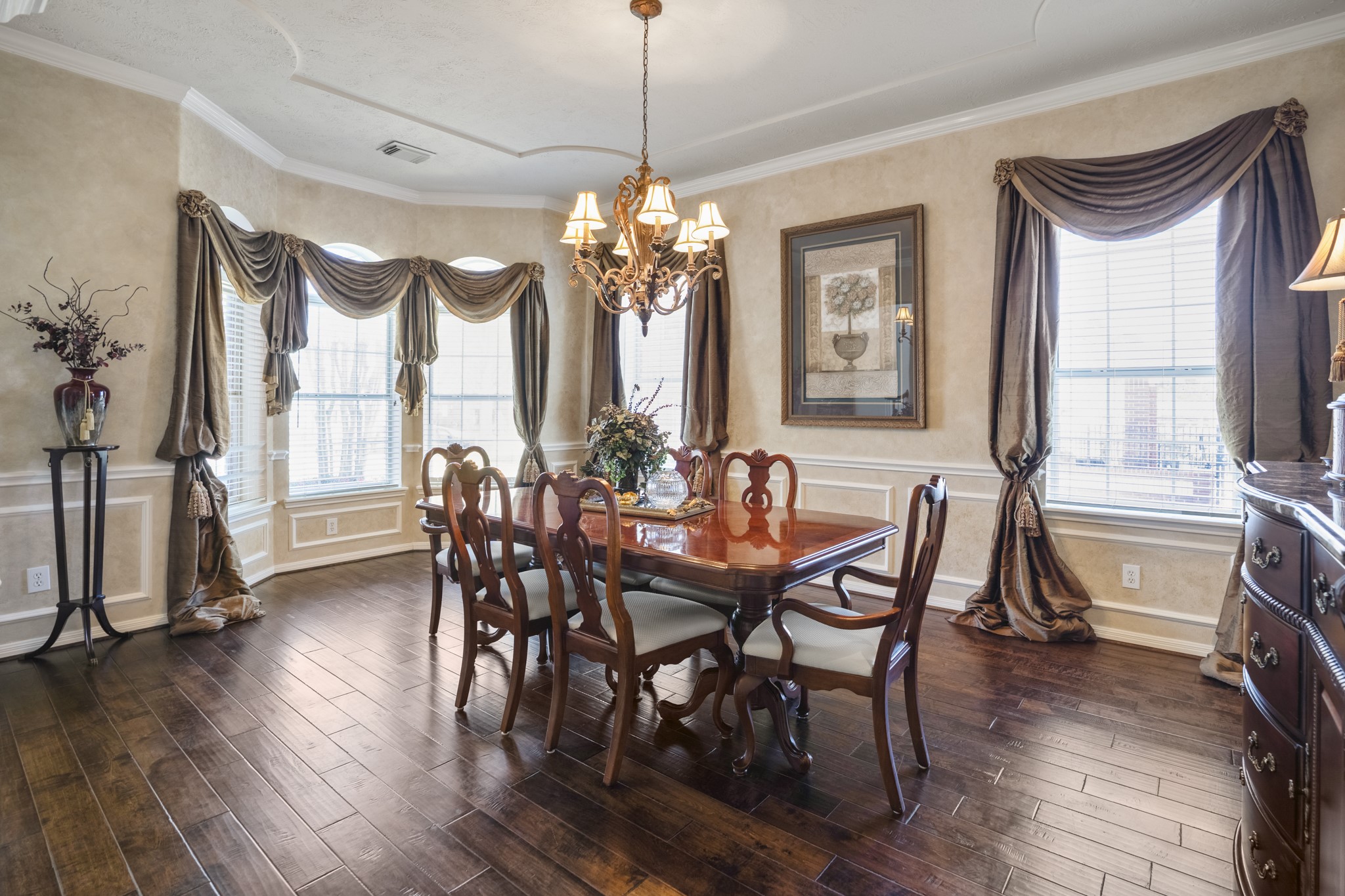 6615 Haughton Drive Spring, TX 77389 - Photo 6 of 50 Inside the formal dining room, the craftsmanship becomes even more apparent. Rich wood flooring grounds the space, while chair railing and box panel trim add depth and character along the walls. Crown moulding frames the room beautifully, drawing your eye up to the scalloped ceiling detail where a grand chandelier anchors the space.
The bay windows bring in soft natural light, balancing the scale so the room feels both impressive and inviting — ready to host gatherings of any size without ever feeling fo