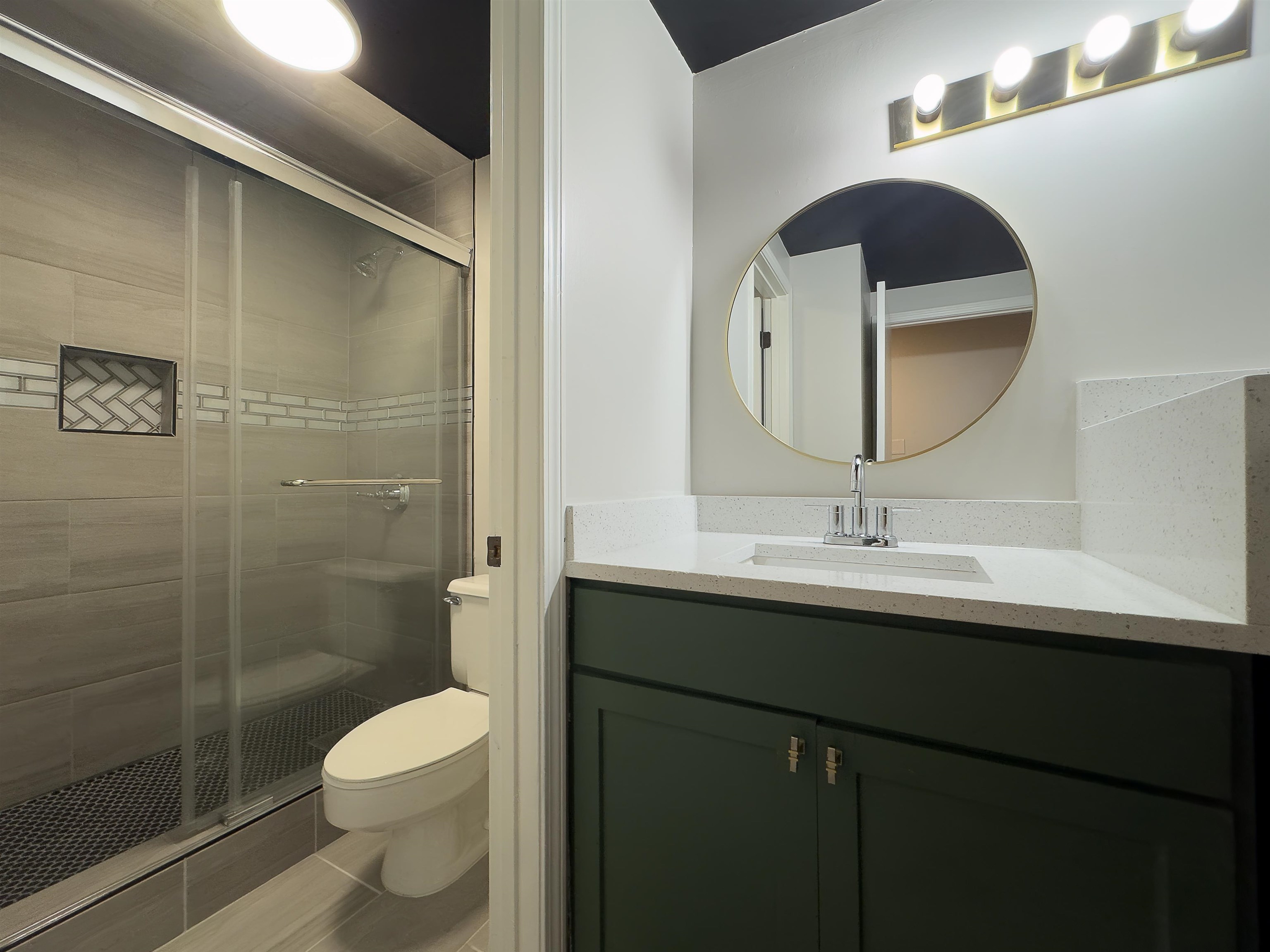 109 North Main Street, Unit 907 Memphis, TN 38103 - Photo 12 of 29 Bathroom featuring a frameless glass shower enclosure, a recessed shower niche with herringbone tile, a dark green vanity with a light-toned countertop, a circular gold-framed mirror, and a four-bulb vanity light fixture
