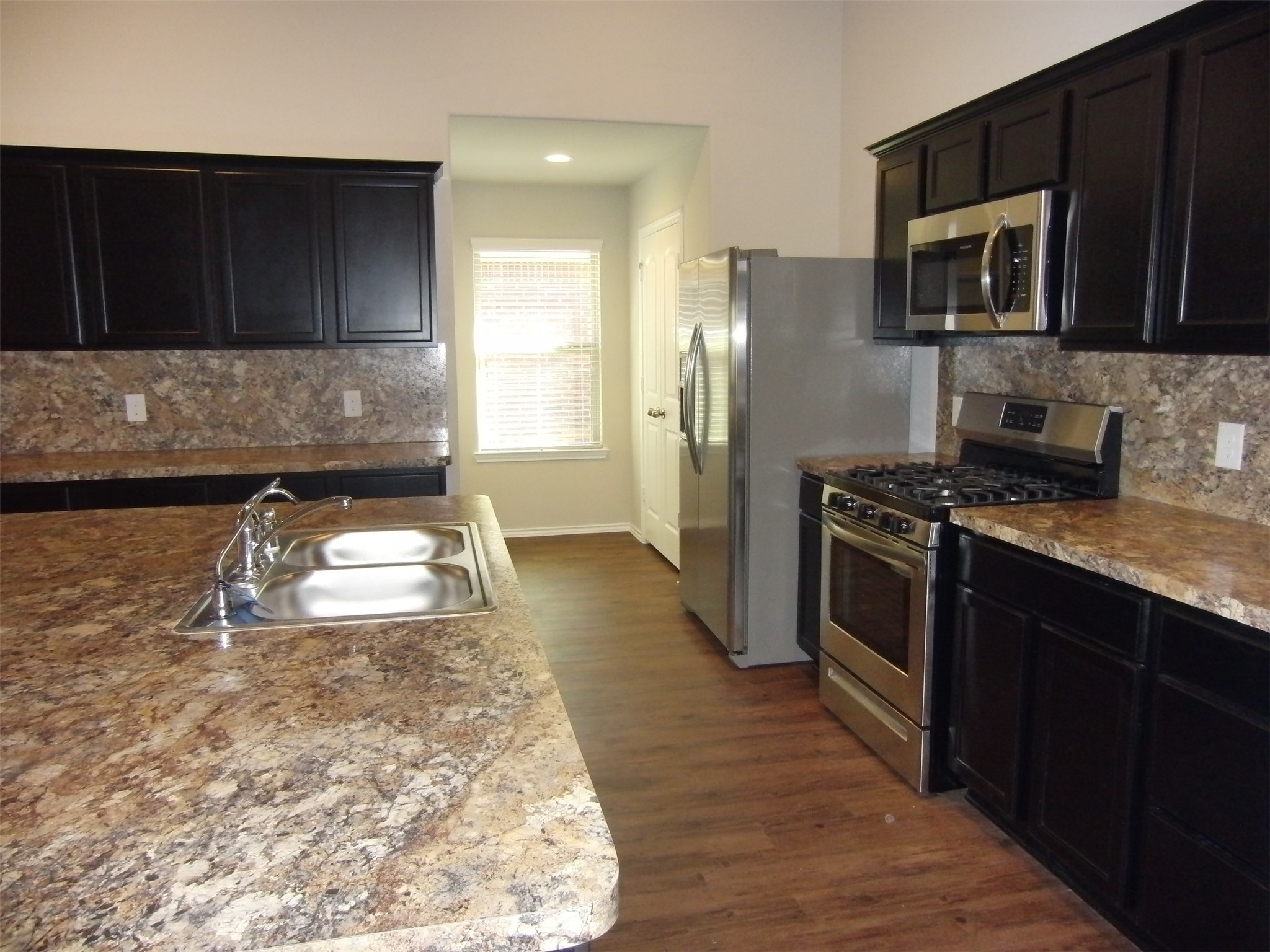 5522 Mesquite Oaks Trail Spring, TX 77389 - Photo 4 of 28 Kitchen features dark cabinetry, stainless steel appliances, and a spacious island with a double sink. The countertops and backsplash have a marble-like finish, complementing the wood flooring and providing a sleek, contemporary look.
