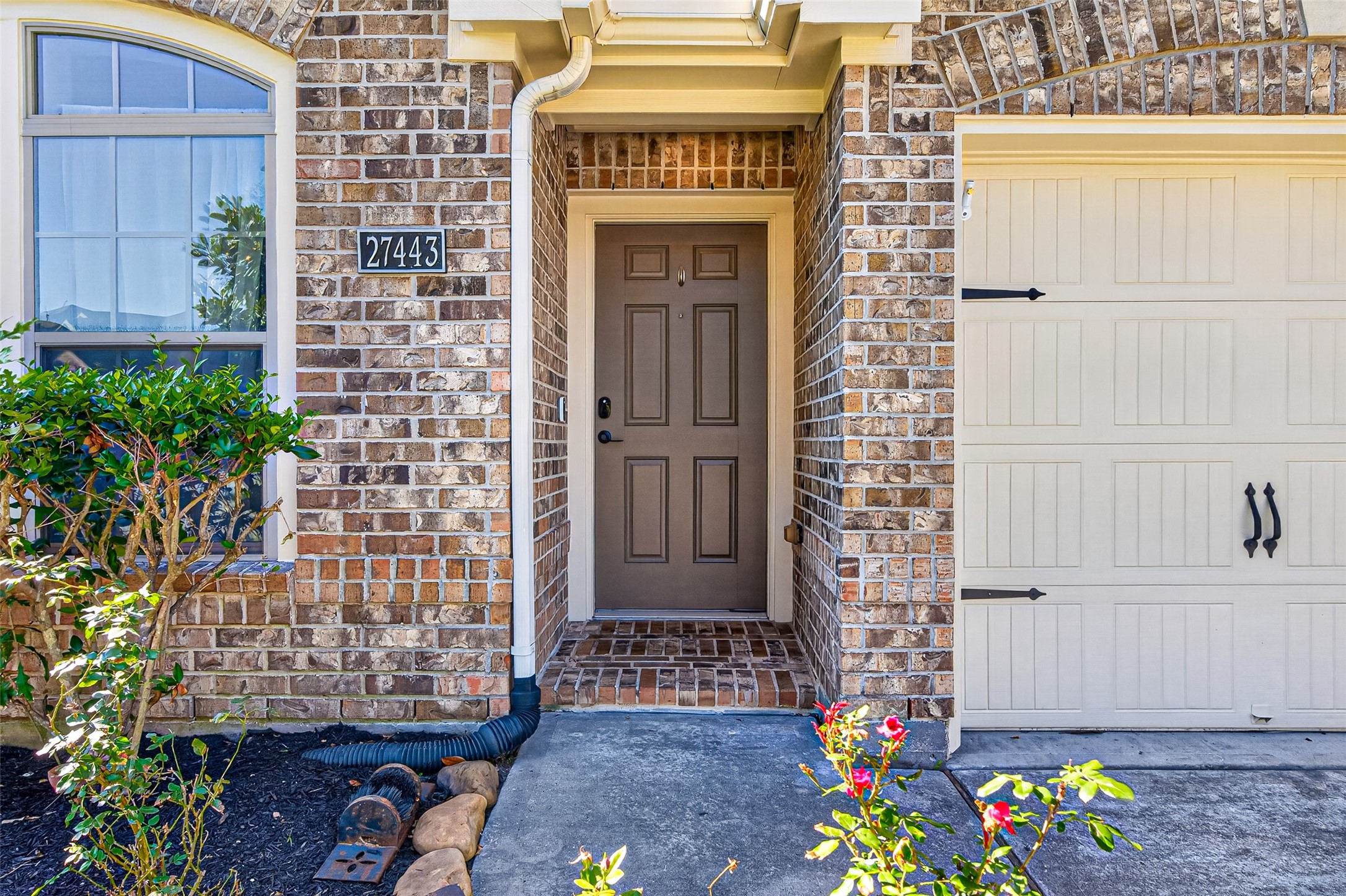 27443 Rebecca Field Lane Spring, TX 77386 - Photo 3 of 31 This photo showcases the inviting entrance of a brick home, featuring a brown front door with a covered entryway. Adjacent is a cream colored garage door. The facade is accented with a well maintained garden and arched window, adding charm and curb appeal.